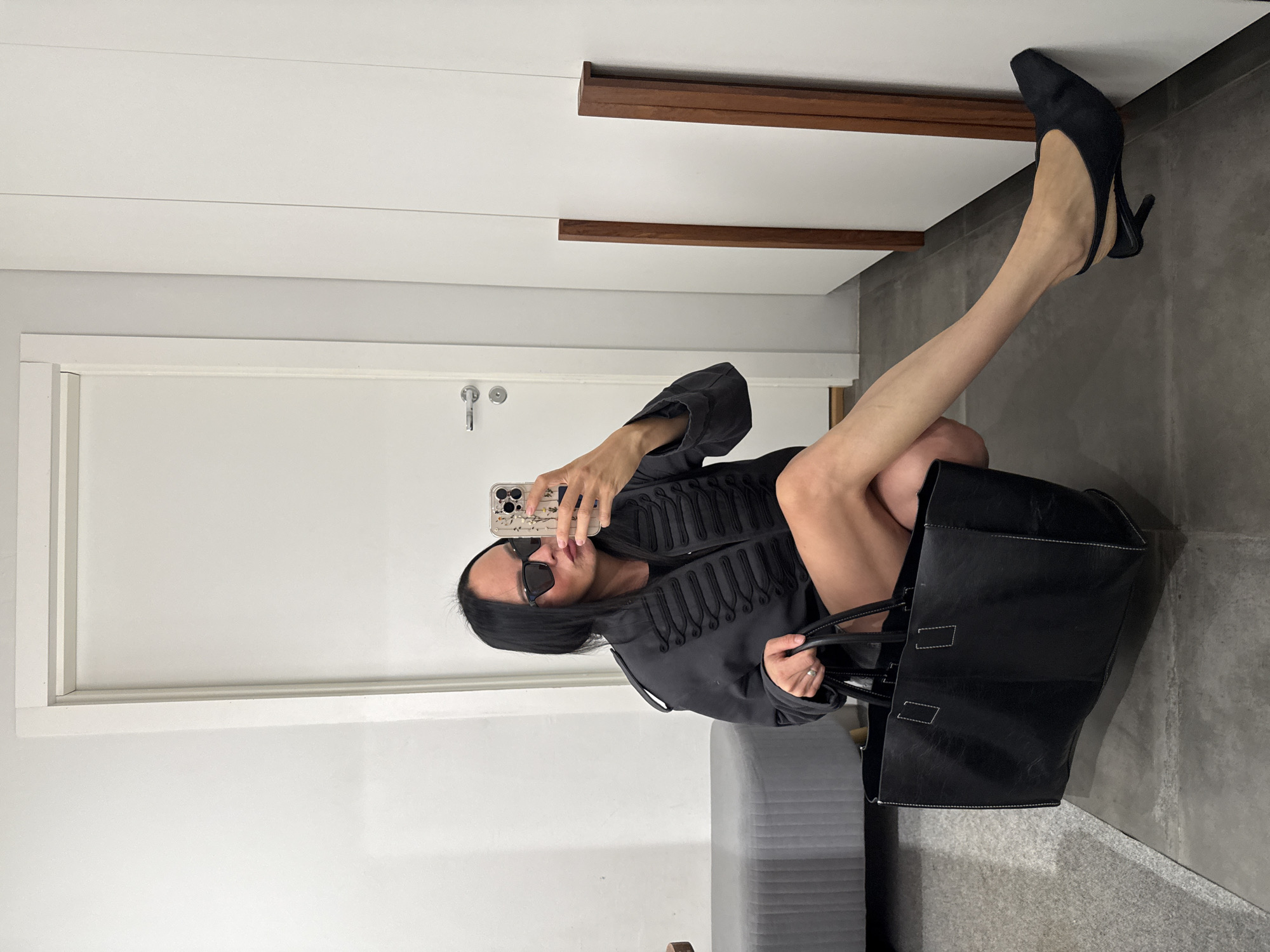 A woman with long dark hair and sunglasses is taking a mirror selfie. She is wearing a dark jacket with ornate braiding and a black dress. She is holding a large black tote bag in her left hand and her right hand is holding her phone. She is crouching down and her legs are crossed. She is wearing black slingback heels. The background appears to be an indoor space, possibly a dressing room or a retail store. There is a white door on the left and white closets or panels on the right. A gray upholstered bench is visible behind her. There is no indication of the city of Chaikovsky, Russia.