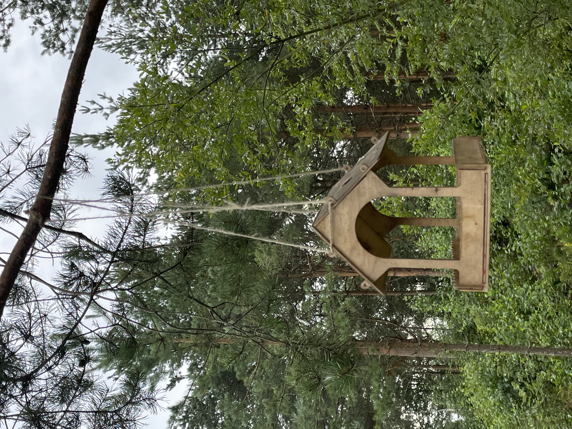 A wooden bird feeder hangs from a tree branch amidst lush green foliage. The feeder has a tiered roof with arched openings on the sides. The surrounding environment appears to be a forest or wooded area with tall trees and dense undergrowth. The lighting suggests an overcast day. The location is identified as Озон Снт, Russia. There are no people or animals visible in the image.