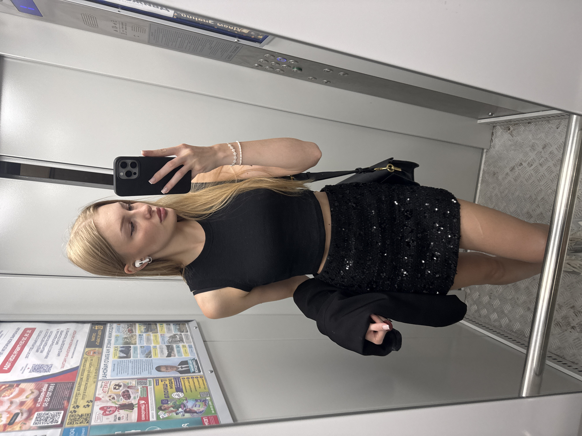 A young Caucasian woman with blonde hair tied in a ponytail is taking a mirror selfie in an elevator. She is wearing a black sleeveless crop top, a black sequined mini-skirt, and white wireless earbuds. She is holding a black smartphone in her right hand and a dark jacket is draped over her left arm. A black purse with a gold clasp is slung over her right shoulder. The elevator has metal walls and a textured metal floor. Behind her, on the left wall, are several posters in Russian. One poster features a man in a suit, likely a political advertisement, and another displays various products and services, including what appears to be a promotion for a local market or sale. The overall environment suggests an urban setting in Perm, Russia, given the Cyrillic text on the posters.