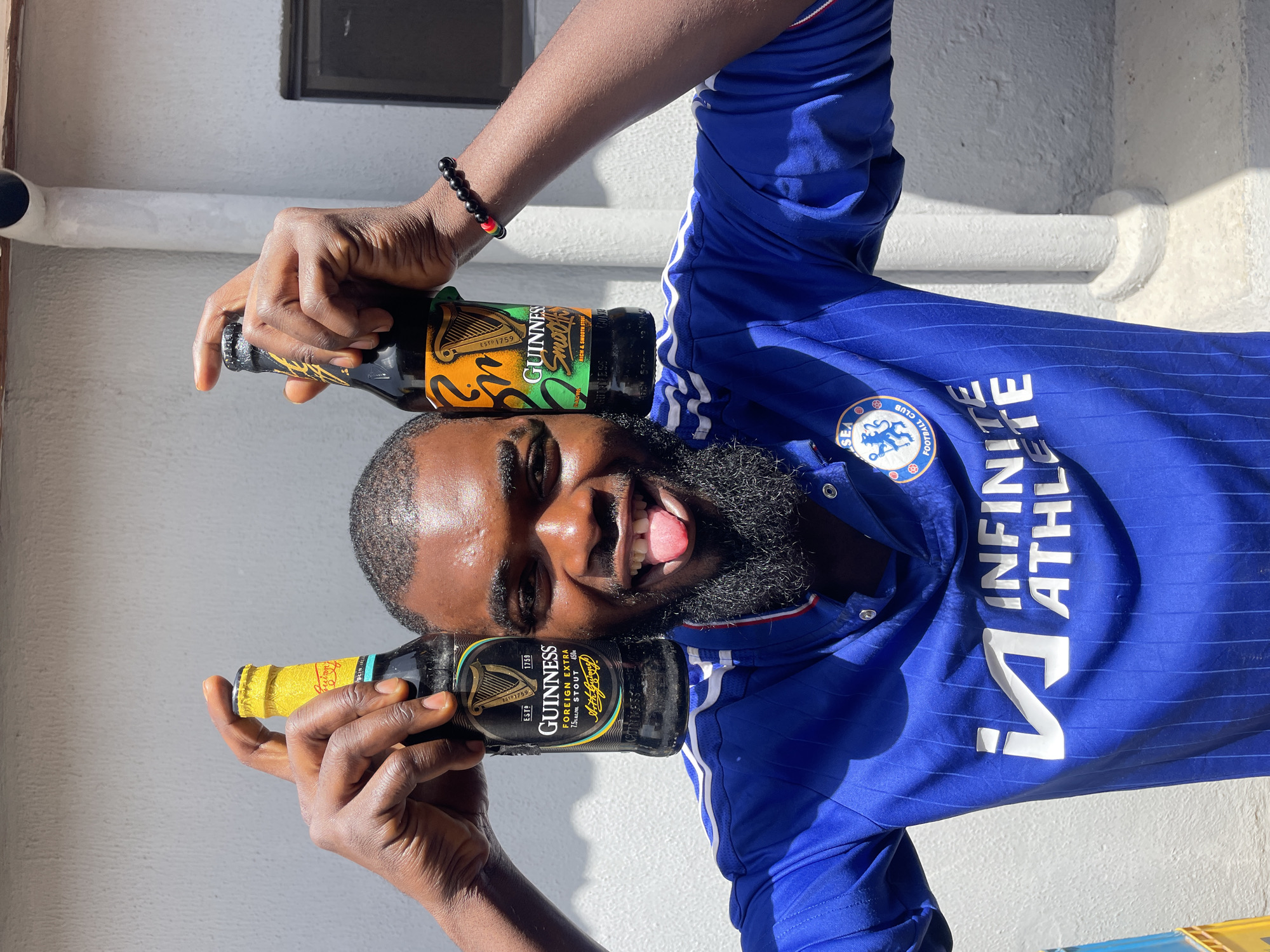 A person with dark skin, a beard, and a bright smile is holding two bottles of Guinness. The person is wearing a blue jersey with white stripes and the Chelsea Football Club logo. The bottles are positioned on either side of the person's head, with one bottle resting on their forehead and the other near their mouth. The Guinness bottles are dark glass with distinctive labels. The person has a red and black beaded bracelet on their wrist. The background appears to be an outdoor setting with a white textured wall and some sunlight casting shadows, suggesting it is daytime. The overall mood is joyful and celebratory, with the person's open mouth and sticking-out tongue conveying amusement. The scene is shot from a low angle, emphasizing the person and the bottles. Visible text on the bottles includes "GUINNESS", "FOREIGN EXTRA STOUT", and "ESTD 1759". The jersey also has text that appears to be "INFINITE ATHLETIC". The scene suggests a moment of lighthearted enjoyment, possibly a break or a celebration.