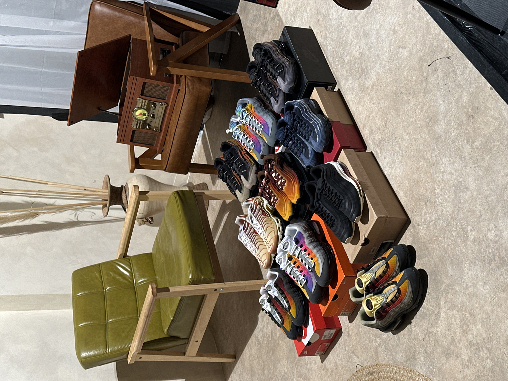 The image shows an indoor scene with a collection of objects arranged on the floor, including two armchairs, a radio, several shoe boxes, and multiple pairs of Nike Air Max 95 sneakers. One armchair is brown leather, while the other has a green padded seat and wooden frame. A vintage-style radio sits near the brown armchair. The shoe boxes, both with and without shoes, are stacked in a disorganized pile. The sneakers vary in color, including black, orange, grey and multicoloured. The image is shot from a high angle, looking down at the items, under bright natural light. There is no evidence indicating the location of this image in London, United Kingdom.