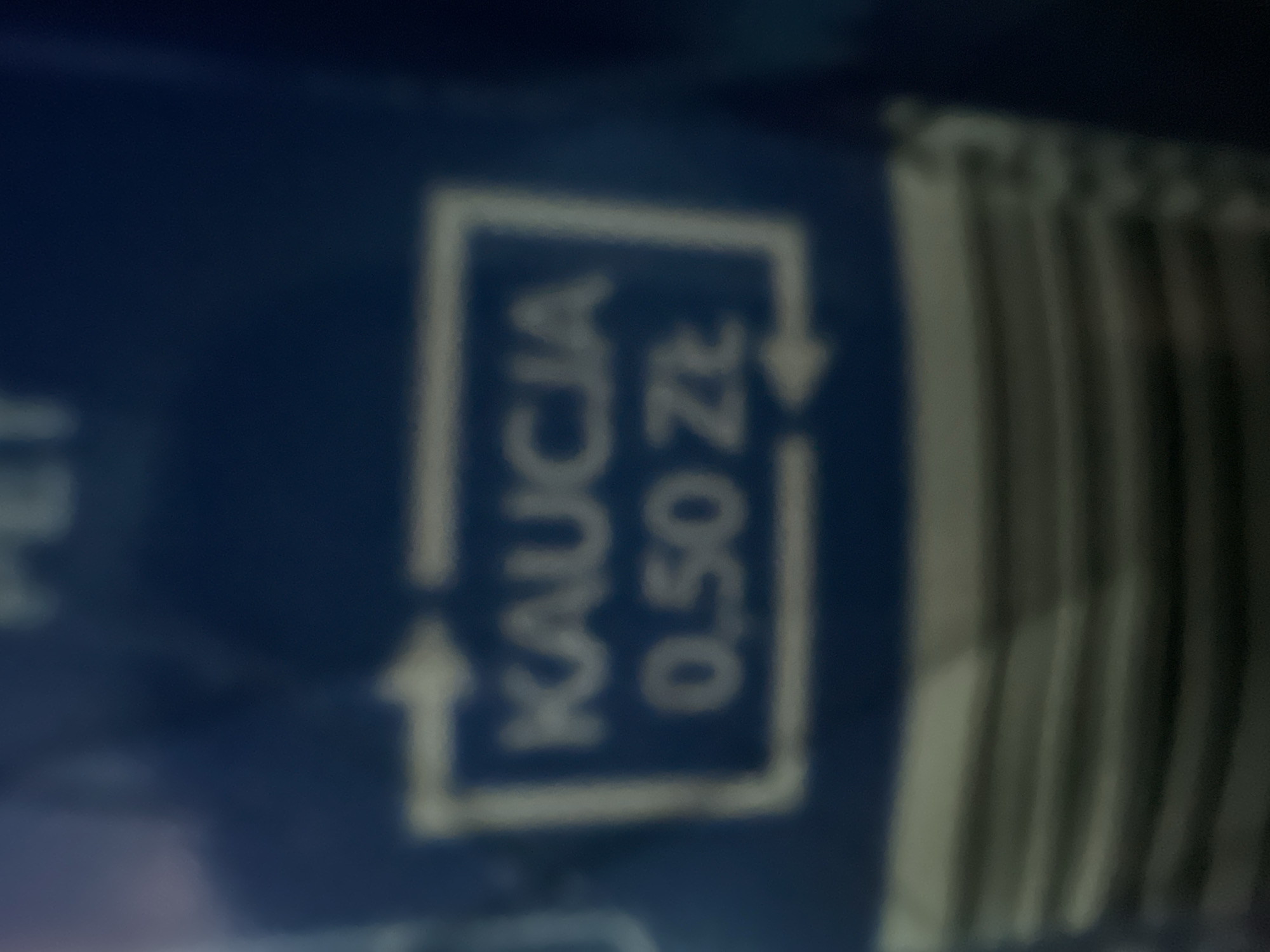 This image shows a close-up, blurry, vertical shot of a dark blue surface, likely a product label or packaging. On the surface, within a white rectangular outline, is white text that reads "KAUCJA 0,50 ZŁ". An arrow points from the left into the rectangle, and another arrow curves out from the right, suggesting a recycling or deposit return symbol. To the right of the symbol, there is a series of vertical lines, possibly indicating a barcode or ribbed texture. The text is in Polish, with "KAUCJA" meaning "deposit" and "0,50 ZŁ" indicating a price of 0.50 Polish Zloty. The lighting is not strong enough to determine the time of day or weather conditions. The setting appears to be indoors, given the focus on the product. There are no people visible.