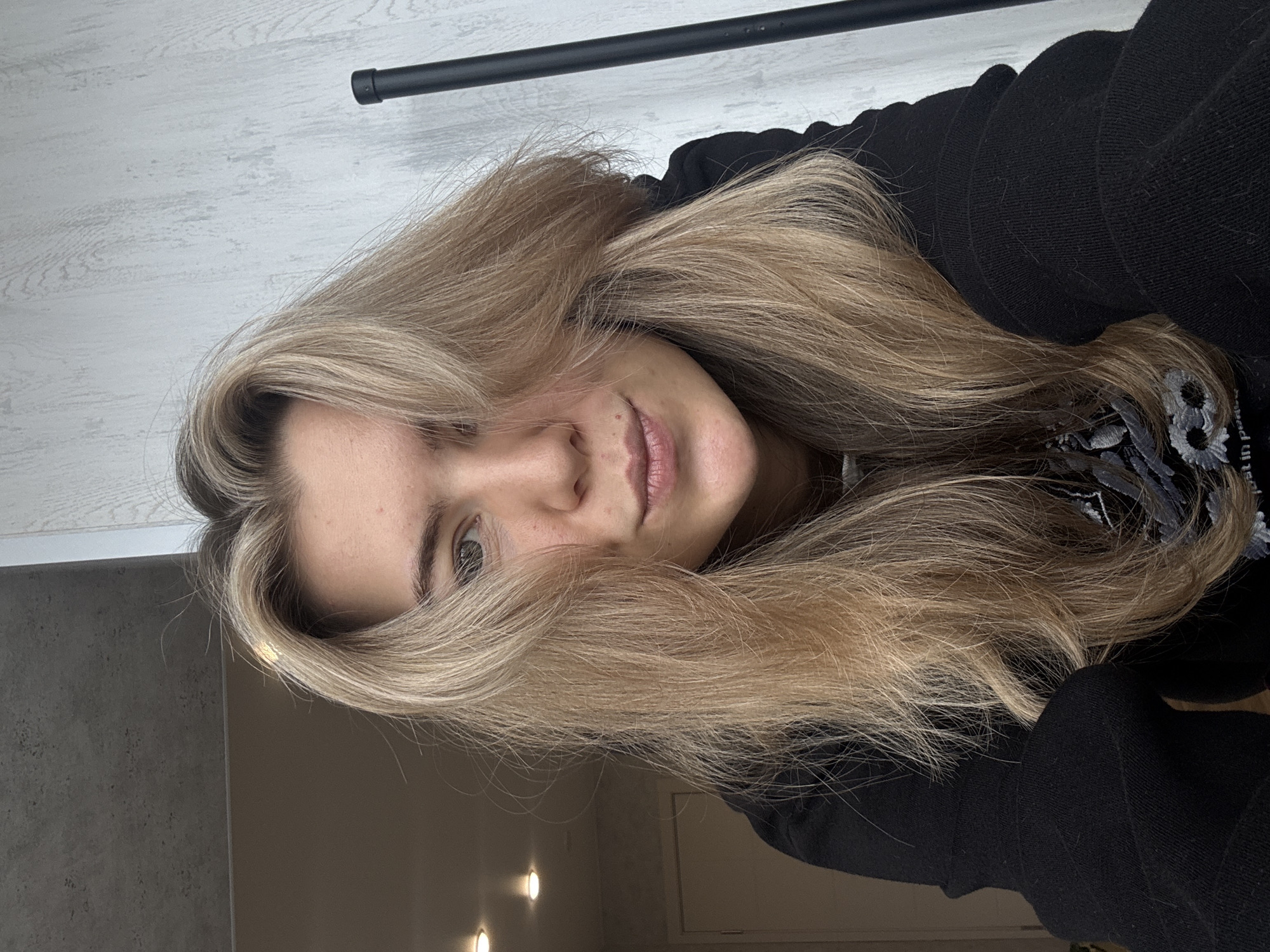 A close-up, rotated image captures a young person lying down and looking towards the camera with a gentle, natural expression. The subject has long, voluminous light blonde hair, streaked with darker roots, which frames their face and partially obscures it. Their left eye is visible, meeting the viewer's gaze.
They are wearing a black long-sleeved top, possibly a hoodie or sweater, with a subtle embroidered or printed pattern in grey and white tones visible on the upper right side. The background behind their head features a light-colored wall with a faint, wood-grain like texture. A dark, horizontal bar or rod stretches across the top of the frame. In the bottom left corner, a darker, plain wall or surface is visible, along with two distinct circular lights, indicating an indoor setting with artificial illumination.
The overall mood of the image is relaxed and casual. A small, partially visible piece of text, "in pe", can be seen within the pattern on the clothing.