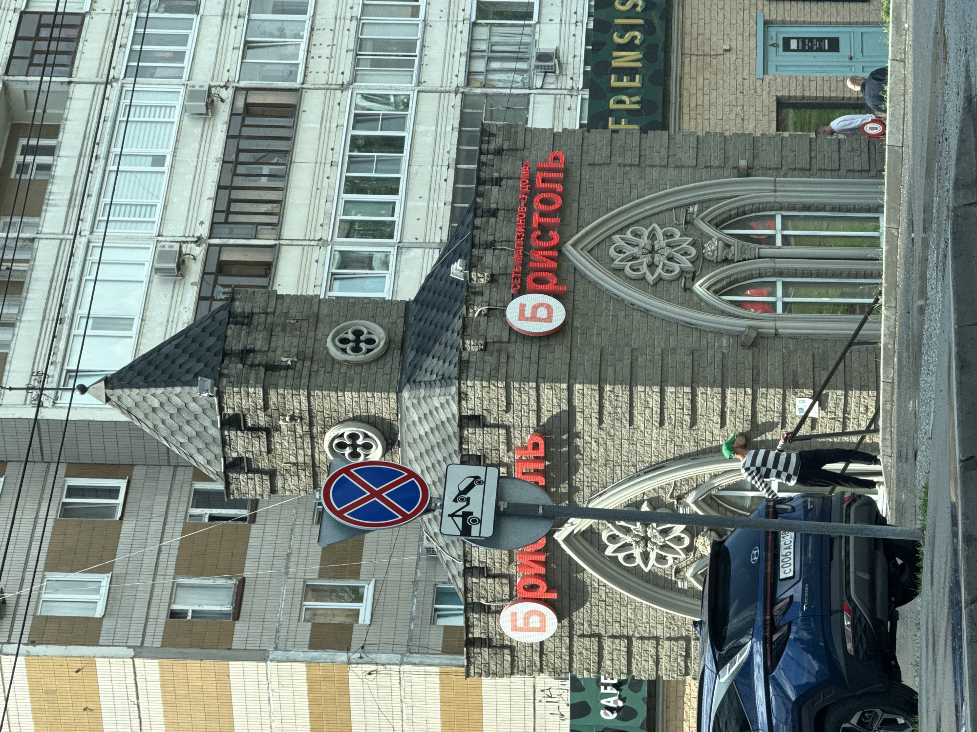The image shows a street scene in Izhevsk, Russia, in front of a building with a distinctive castle-like facade. A blue Hyundai Tucson is parked on the left, and a man in a striped shirt and black pants is standing on steps to the right, holding onto a railing. A tall traffic sign with a "no parking" symbol and a towing symbol is visible in front of the building. The building has a sign that reads "Бристоль" (Bristol) in large red letters, indicating a store. Another sign above it reads "СЕТЬ МАГАЗИНОВ У ДОМА" (Network of neighborhood stores). There are several windows on the building's facade. In the background, apartment buildings with multiple floors and windows are visible. The street in the foreground appears wet, suggesting recent rain.