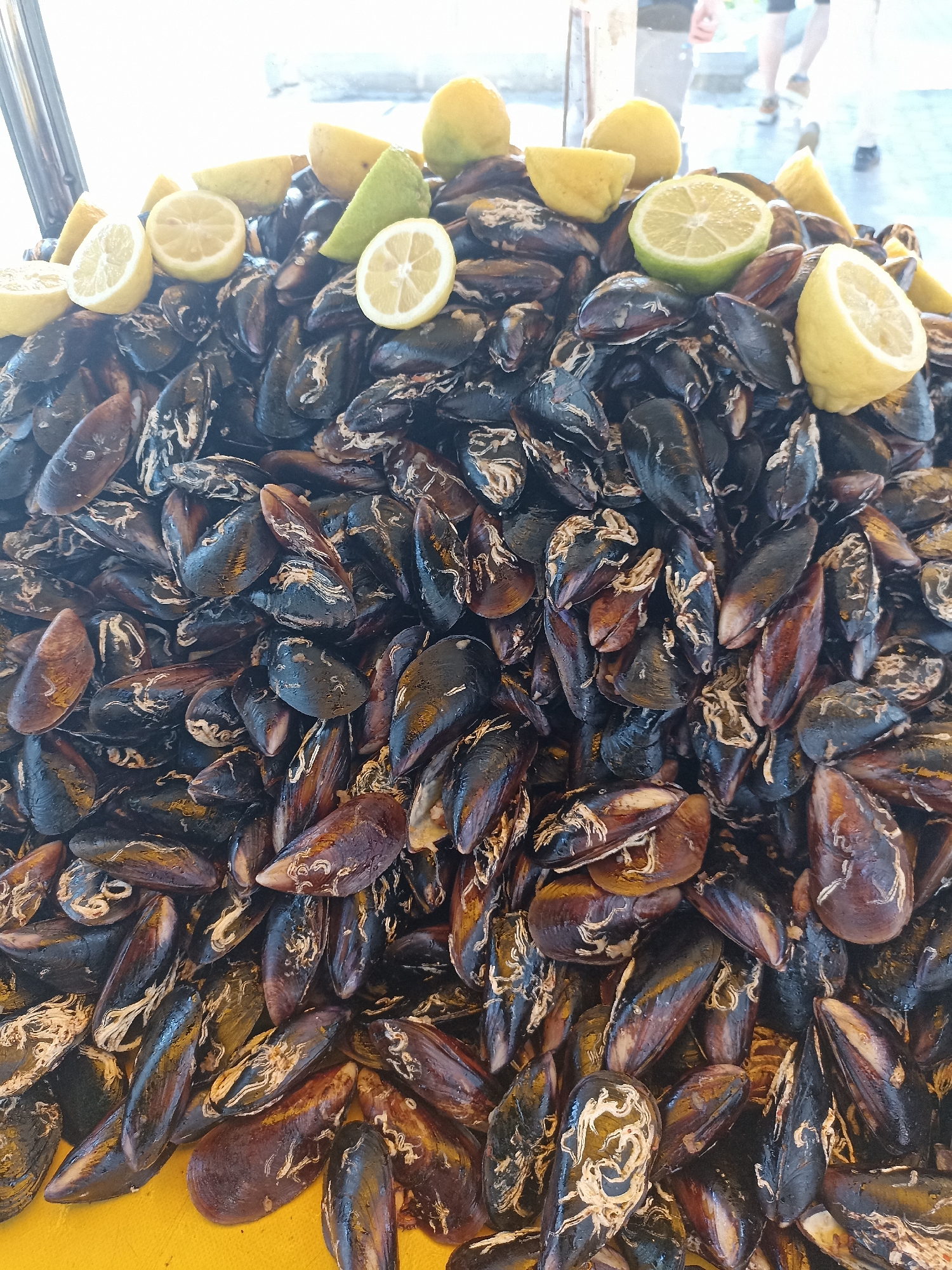 A high-angle shot shows a large pile of dark-colored mussels on a yellow surface, topped with several lemon halves and wedges. The mussels are predominantly black and brown with a glossy sheen. The lemon pieces are yellow and green. In the blurred background, people are walking on a paved surface. The location is Yenişehir, Turkey.