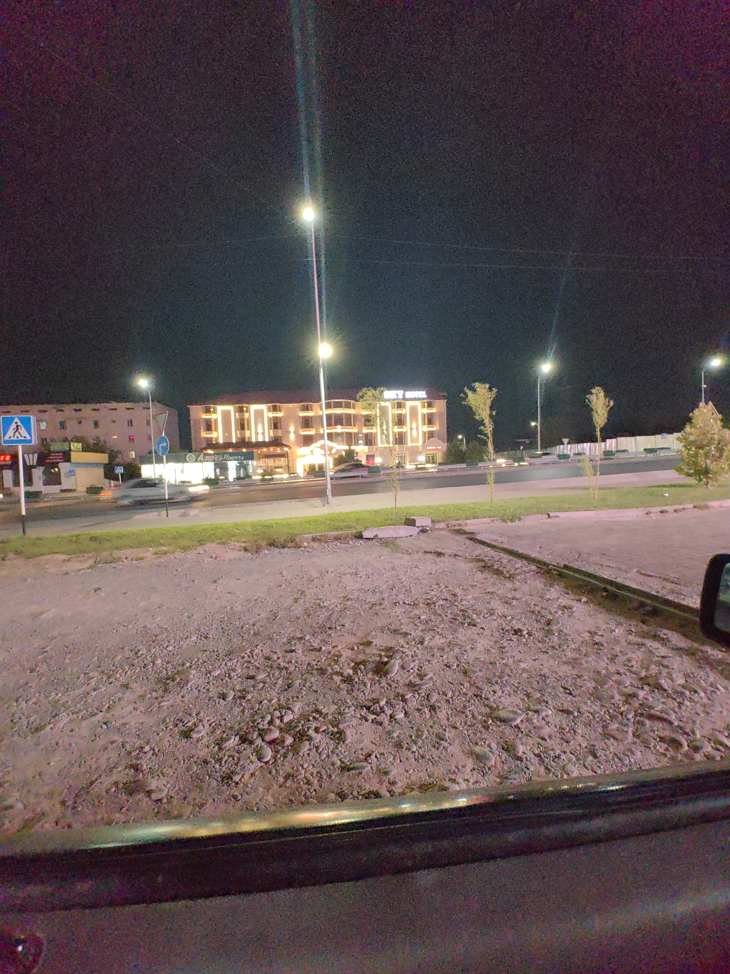 The image depicts a nighttime street scene in the city of Shymkent, Kazakhstan. In the background, a multi-story building with illuminated windows and signage that reads "SKY HOTEL" is visible. To the left of the hotel, another building with a similar architectural style and illumination is present. Streetlights illuminate the road, and trees line the sidewalk. A pedestrian crossing sign and a blue circular traffic sign are also visible. The foreground consists of a gravelly ground surface, likely a roadside or parking area, with a dark vehicle's interior visible in the lower portion of the image. The overall atmosphere suggests a commercial or residential area in an urban setting at night.