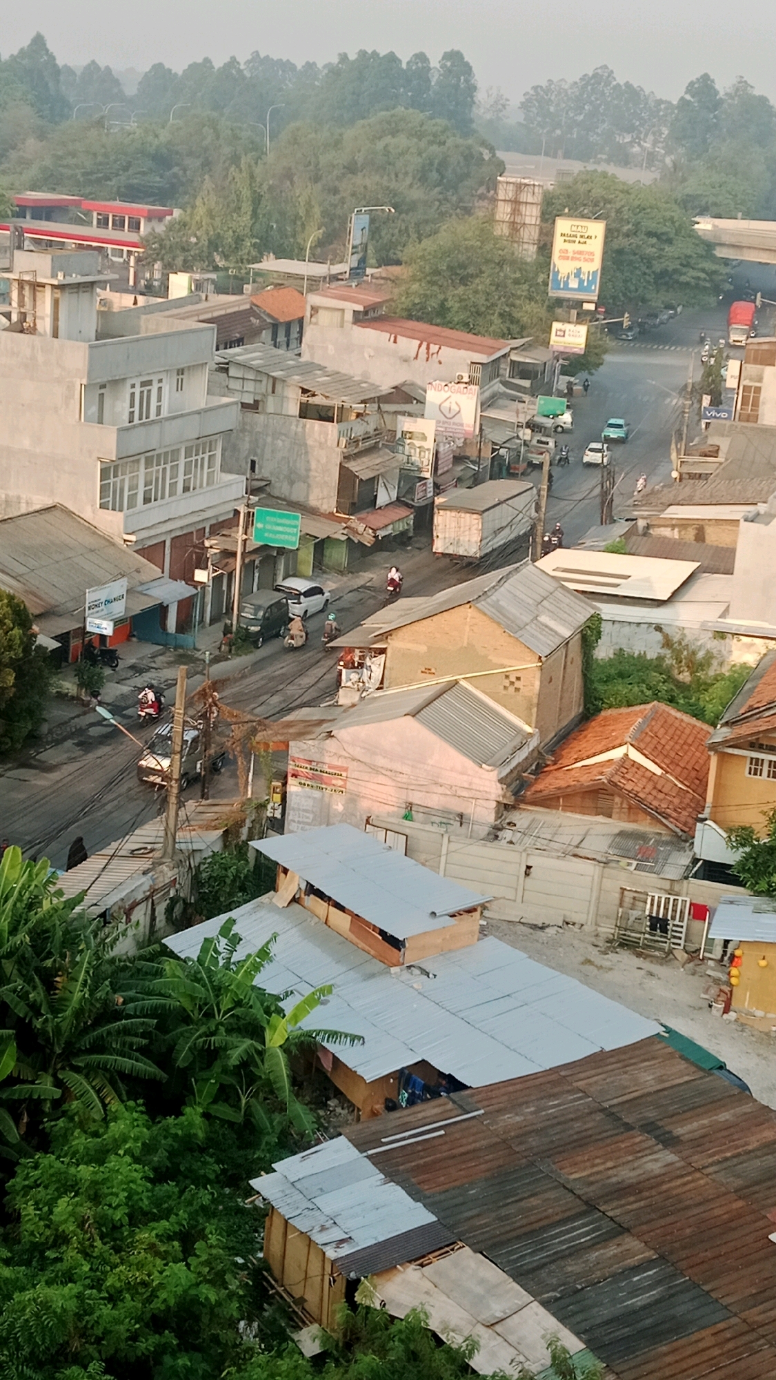 This aerial view captures a dense urban landscape in Tangerang, Indonesia, characterized by a mix of residential and commercial structures, and bustling street activity. The scene is dominated by a network of roads and alleyways winding through a dense collection of buildings with a variety of roofing materials, including corrugated metal, tiles, and flat surfaces.
In the foreground, several residential structures are visible, some with corrugated metal roofs and others with lighter-colored flat roofs. Green foliage, including what appear to be banana trees, is interspersed among the buildings, suggesting a humid climate.
Further into the image, commercial buildings are more prominent. A multi-story white building with numerous windows stands out on the left. Across the street, a row of smaller, single-story shops and houses with varied facades line the road.
The streets are active with vehicular traffic, primarily motorcycles and a few cars. Several people are seen riding motorcycles. A white van is parked on the side of the road. A larger, box-shaped truck is visible further down the street.
Signs are present throughout the scene, some displaying text in what appears to be Indonesian. One prominent sign, possibly an advertisement, features a stylized image of buildings and text. Another sign reads "MONEY CHANGER."
The environment appears to be a typical urban setting with visible electrical wires and poles. The background reveals more trees and buildings, with what seems to be an overpass or elevated road structure in the distance. The overall impression is one of a lively and densely populated area, indicative of a commercial and residential hub within Tangerang.