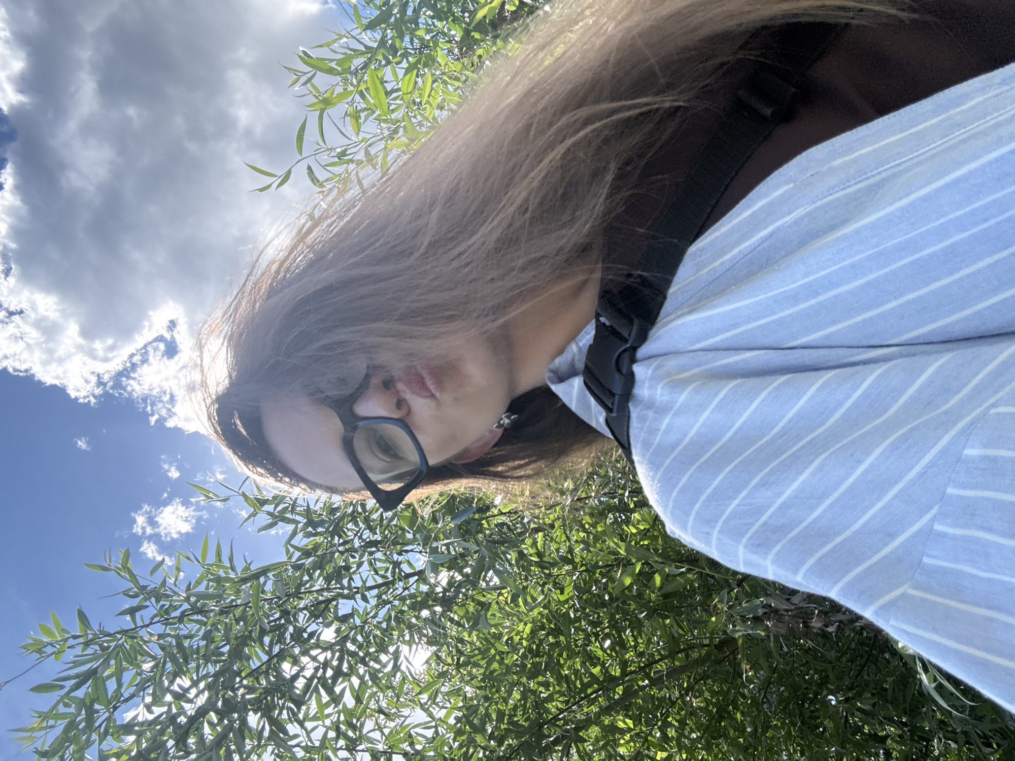 A low-angle, medium shot shows a woman from the chest up, posing for a selfie outdoors. She is wearing dark-rimmed glasses and has long, brown hair that is blowing in the wind. Her lips are pursed. She is wearing a light blue and white striped shirt over a dark brown garment, with a black strap visible across her chest. Behind her, green foliage fills the left and right sides of the frame, with a bright blue sky and white clouds occupying the upper portion. The lighting suggests it is daytime, possibly with the sun behind her. The scene appears to be in a natural outdoor environment, possibly a park or wooded area. The exact location in Chaikovsky, Russia cannot be determined from the image alone.