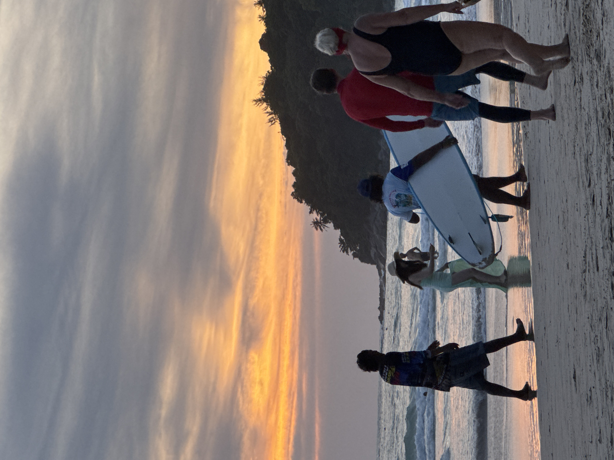 At Karon Beach, Thailand, during sunset, a group of individuals walks along the shoreline. The sky is illuminated with vibrant orange and pink hues from the setting sun, casting a warm glow on the scene. Several people are visible: a person in the foreground is seen from behind, wearing a colorful patterned shirt and shorts. To their right, a woman in a light-colored dress walks, holding what appears to be a beach towel. Further to the right, two individuals are walking together. One is wearing a red long-sleeved shirt and blue shorts, while the other, a woman, is in a black swimsuit. A man carrying a surfboard walks in the middle ground, facing away from the camera. In the background, gentle waves lap the sandy beach, and a lush, tree-covered hill rises from the coastline, silhouetted against the dramatic sky. The overall atmosphere is serene and tranquil, capturing a moment of leisure at the beach as day transitions into night.