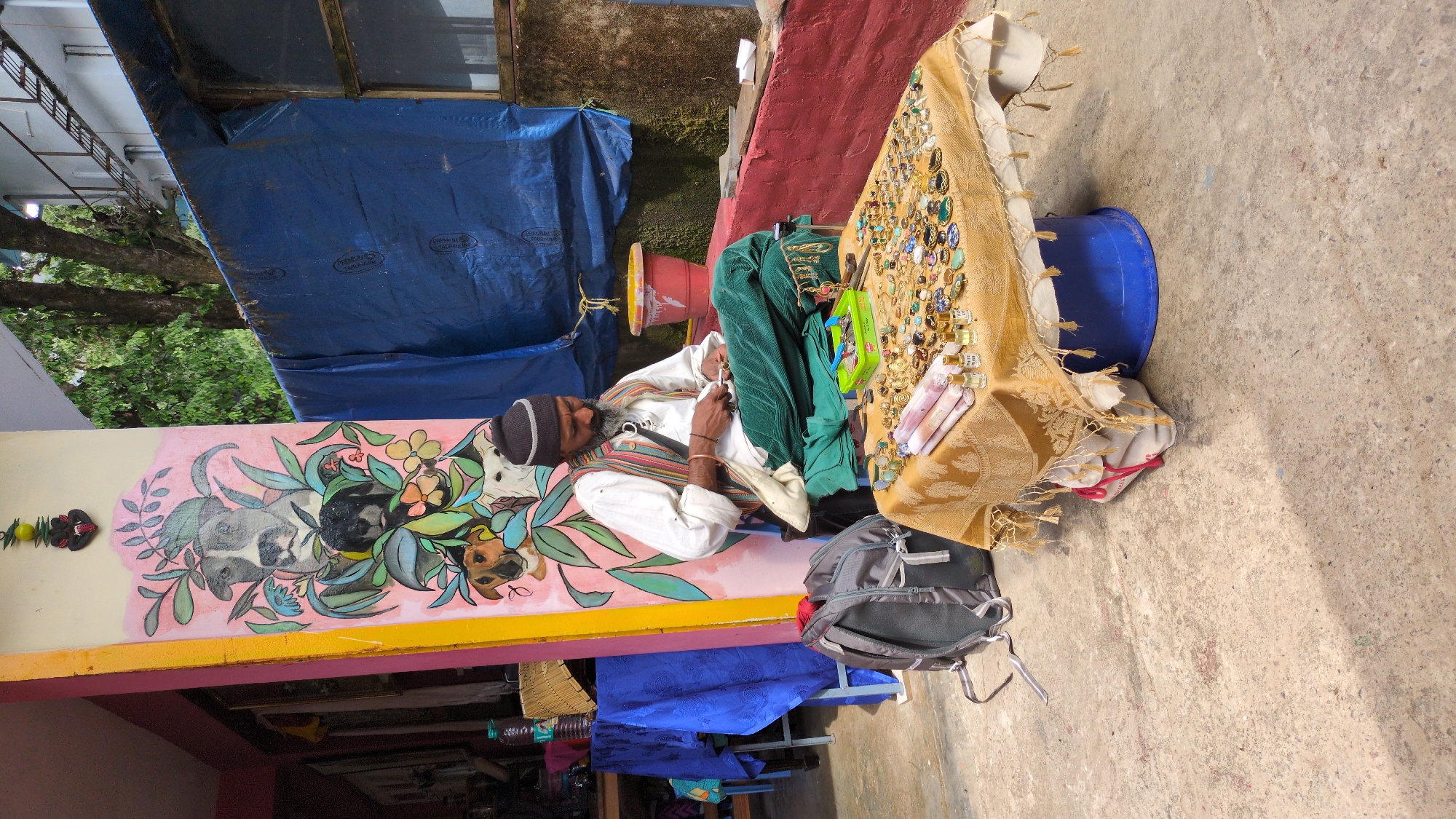The image depicts an outdoor market scene in Bhagsu, India, under bright daylight. An older man, appearing to be a vendor, is seated by a stall, seemingly resting or looking down. He has a dark beard and wears a black beanie, a white long-sleeved shirt, and a colorful striped vest or shawl in hues of brown, green, and red.
The market stall is covered with a golden, patterned fabric, displaying a diverse array of items for sale. These include numerous rings, necklaces, and pendants, many adorned with colorful stones, as well as small clear bottles, likely containing perfumes or essential oils. A folded green garment or fabric rests on the stall next to the man. On the ground beside the stall is a grey backpack with black straps, and a blue bucket is visible beneath the table.
Behind the man, a low wall features a vibrant mural with animal faces, possibly dogs and cats, interwoven with green leaves and colorful flowers on a pink background. Beyond the stall, a section of a red-painted wall is visible, and a large blue tarp is partially seen in the background. The tarp bears faint text, including "PREMIUM" and "TARPAULIN". Green trees and foliage are visible further in the background, suggesting a natural environment. The strong shadows indicate a sunny day.