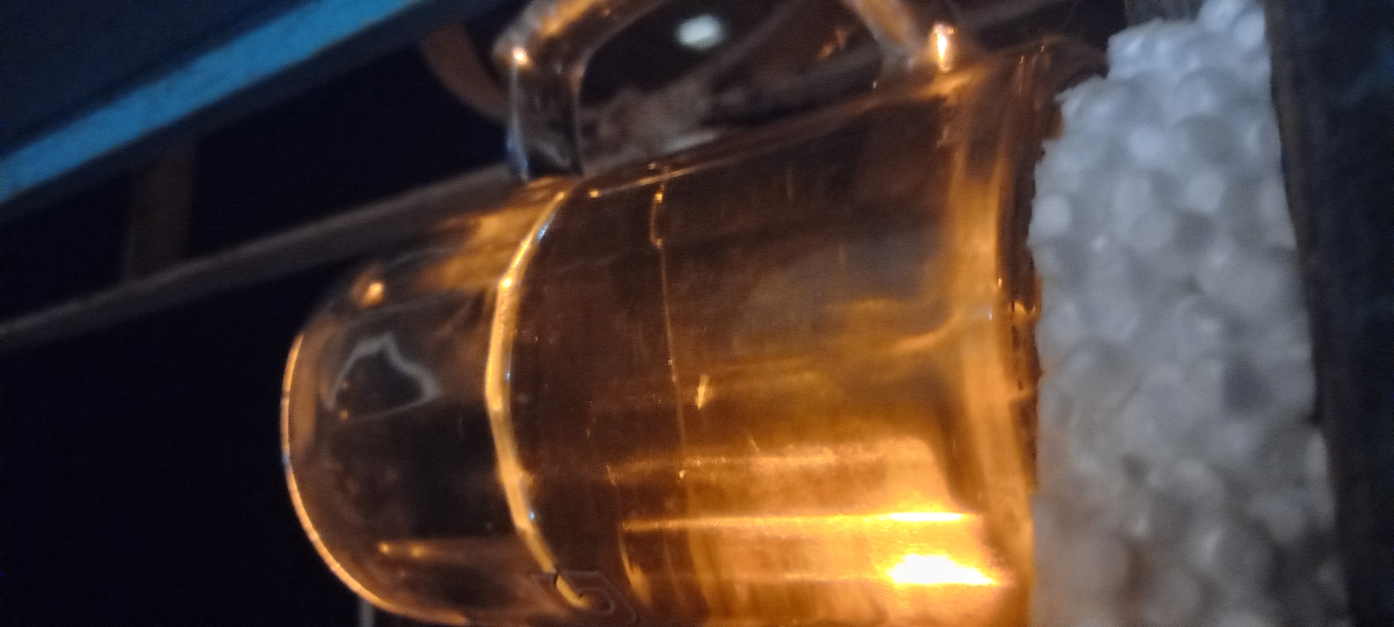 A close-up, low-angle, nighttime shot focuses on a translucent, amber-colored, cylindrical object, possibly a glass or mug, partially filled with a liquid that catches and refracts light. The object rests on a surface covered with small, irregularly shaped, white, granular material, resembling Styrofoam packing peanuts or rice. The background is dark and out of focus, with some indistinct linear elements, suggesting an indoor or sheltered environment. No people, specific landmarks, or details indicating the city of Kryvyi Rih, Ukraine, are visible.