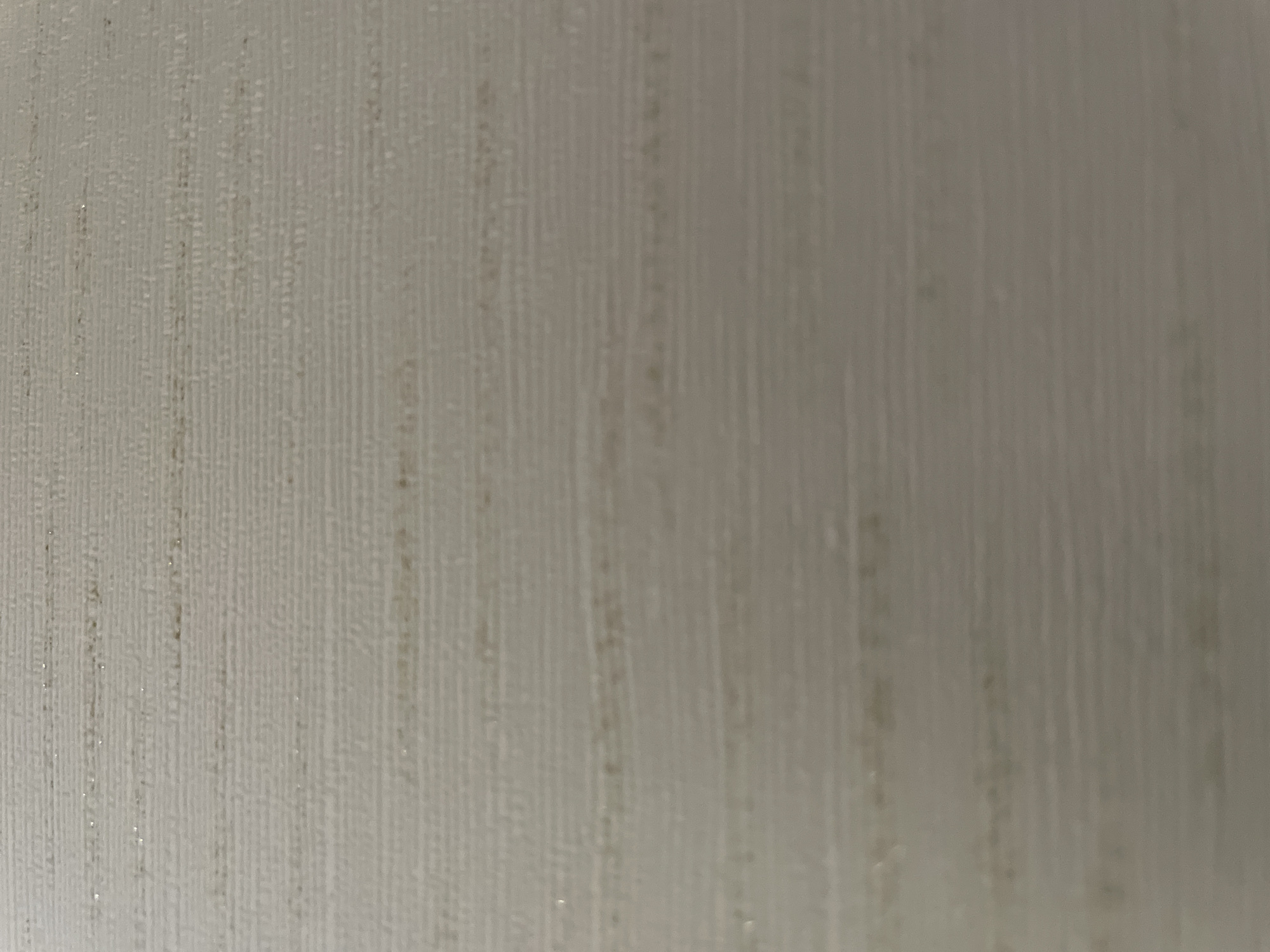 The image shows a close-up, abstract view of a textured surface, likely wallpaper or a similar wall covering. The surface has a subtle horizontal ribbed pattern with faint, irregular darker lines and occasional shimmering flecks, possibly metallic threads. The overall color is a muted, light beige or off-white, gradually darkening towards the bottom of the frame. The texture appears slightly uneven, with small, raised or indented areas contributing to the textured effect. There are no discernible objects, people, or specific environmental features that would allow for localization within the city of Omsk, Russia, or any other location.