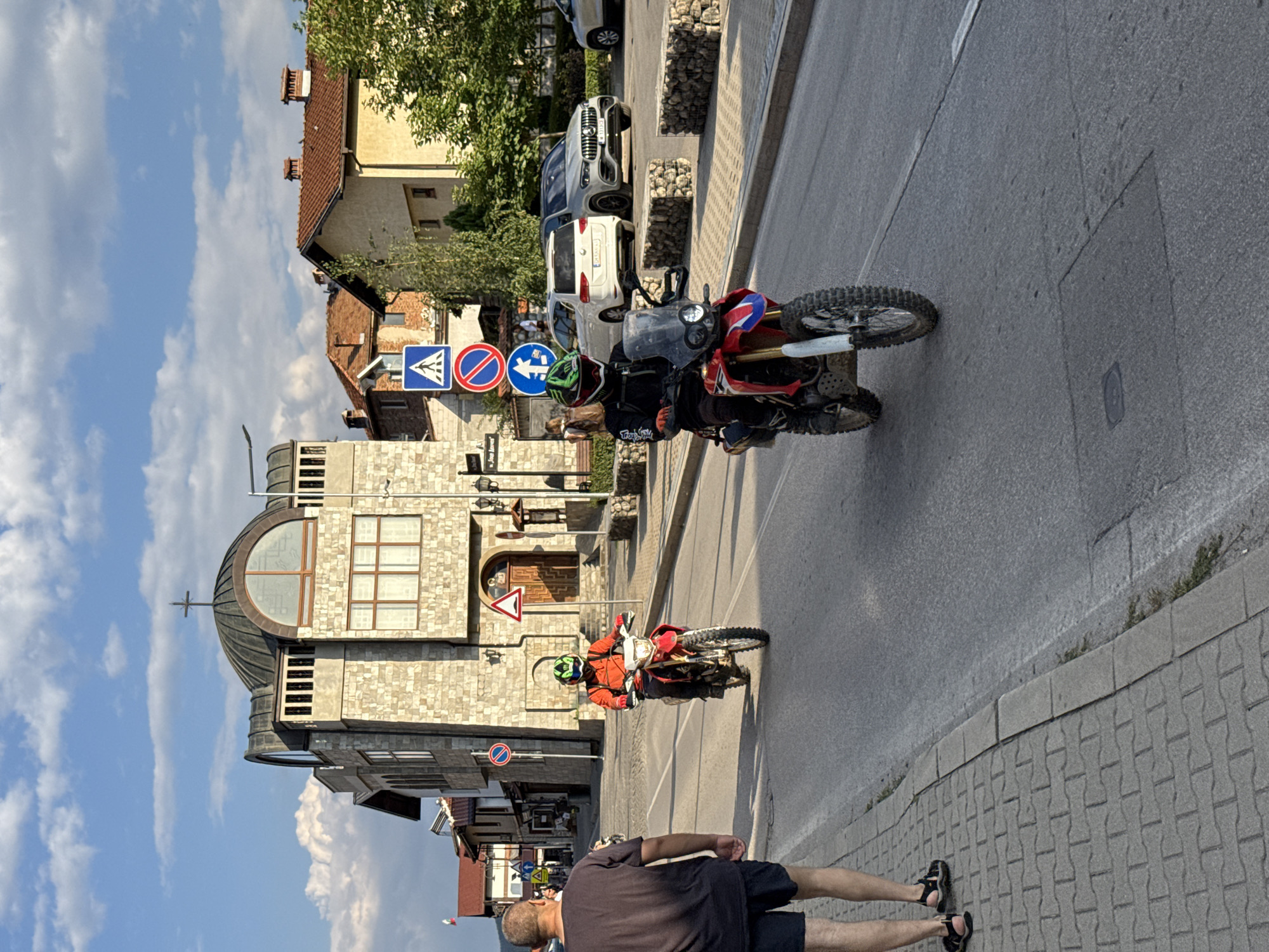 In Bansko, Bulgaria, on a paved street bordered by a brick sidewalk, two individuals on dirt bikes are captured in motion. The person in the foreground, wearing a black and white patterned jacket, green helmet, and black pants, rides a red and white dirt bike. Another individual, in an orange jacket and green helmet, on a red dirt bike, is further up the street. A man in a dark t-shirt and shorts walks on the sidewalk, facing away from the camera. Behind them, a stone building with a domed roof topped by a cross stands prominently, characteristic of religious architecture. Several cars are parked on the right side of the street. Traffic signs, including a pedestrian crossing and a no-entry sign, are visible. The sky above is a clear blue with scattered white clouds. The scene is illuminated by natural sunlight, suggesting it is daytime.