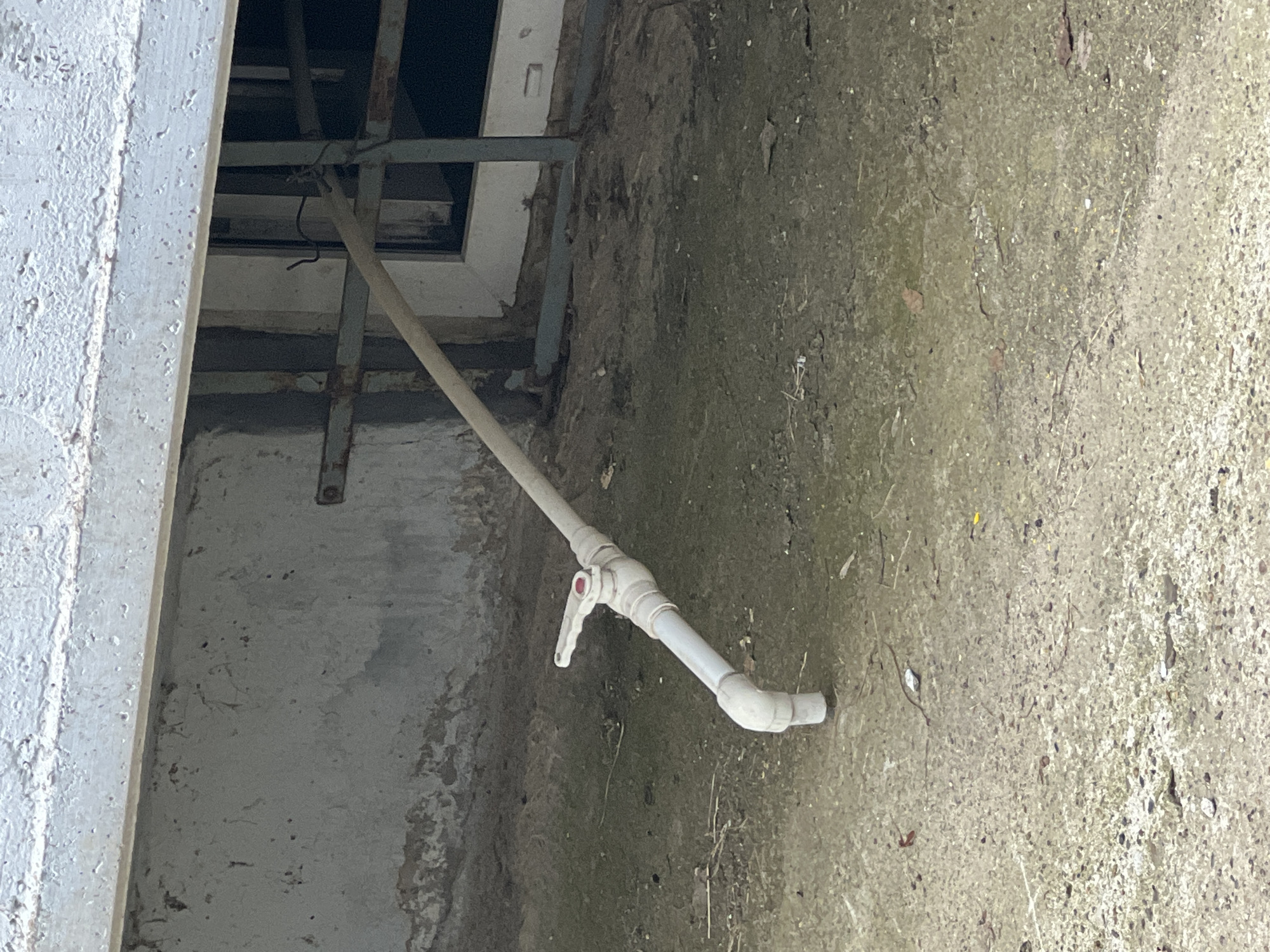 The image displays a ground-level view of a utilitarian outdoor or semi-enclosed area, characterized by rough concrete surfaces. The main subject is a light-colored, possibly white PVC pipe, extending diagonally from the upper left towards the lower right. This pipe appears to emerge from a dark, recessed opening in the background, which is framed by metal bars or supports and a white-painted edge.
Approximately halfway along its visible length, the pipe features a white, quarter-turn valve with a red indicator dot, suggesting a control point for fluid flow. The pipe continues past the valve, ending in an elbow joint that directs its opening close to the ground.
The ground surface is a mix of weathered concrete and packed dirt, with scattered small stones, dry leaves, and other debris. To the far left, a textured, white-painted concrete wall or beam runs vertically, showing signs of wear and flaking paint. The overall lighting suggests it is daytime, with some subtle shadows indicating natural light. There is no visible text or identifiable human activity. The scene conveys a sense of a functional or storage area, possibly neglected, typical of an industrial or utility space.