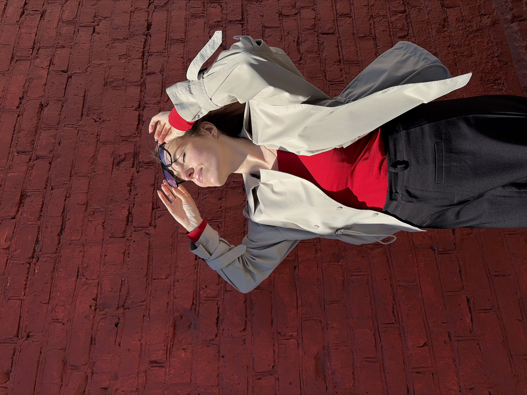 A woman is lying on a red brick surface. She is wearing black pants, a red shirt, and a beige trench coat. Her glasses are in her hand near her forehead. The location may be in the city of Izhevsk, Russia.