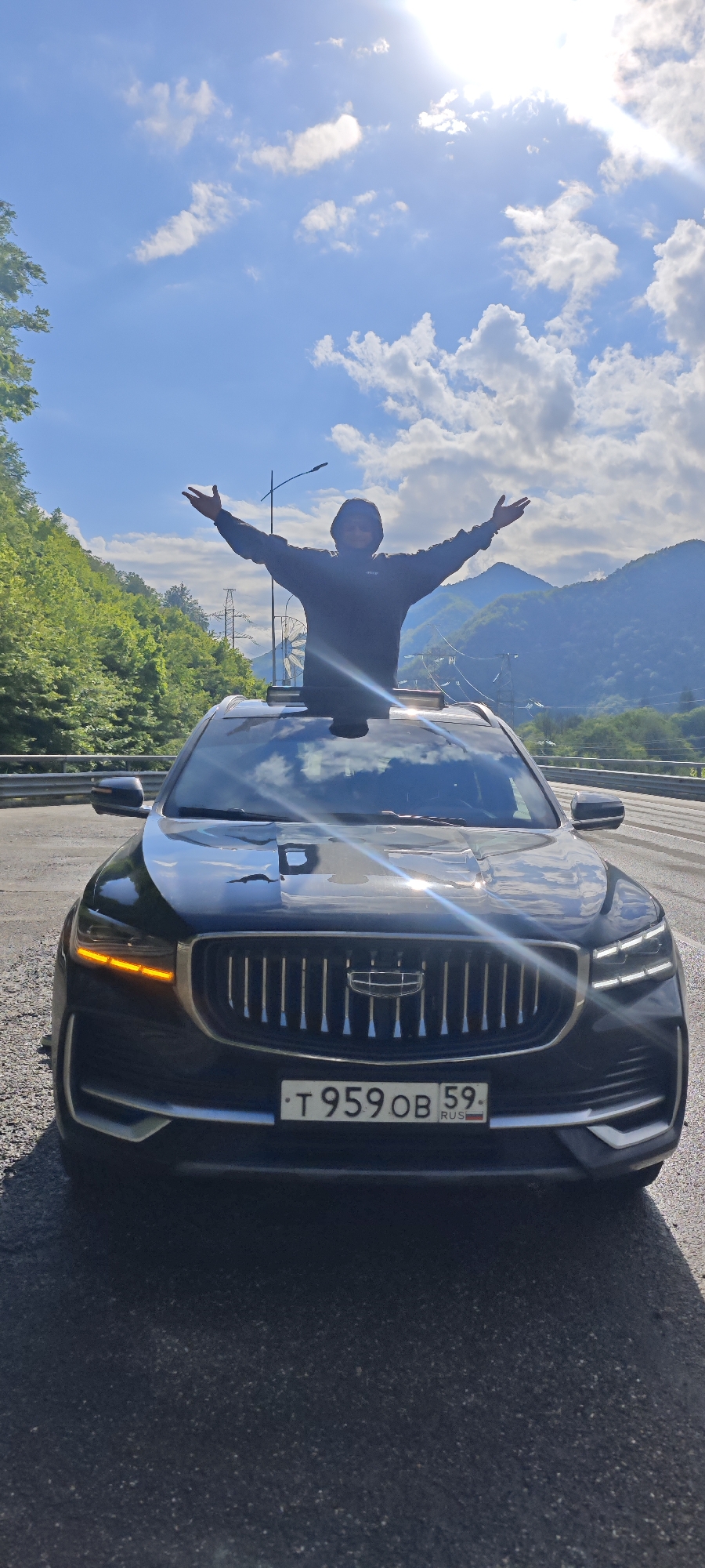 A person stands through the sunroof of a black Geely car, arms outstretched, on a road with mountains and trees in the background. The car has a Russian license plate reading "T959OB 59 RUS." The scene appears to be in a mountainous region, likely near Krasnaya Polyana, Russia, given the landscape and the license plate. The sky is blue with scattered clouds, and the sun is shining brightly.