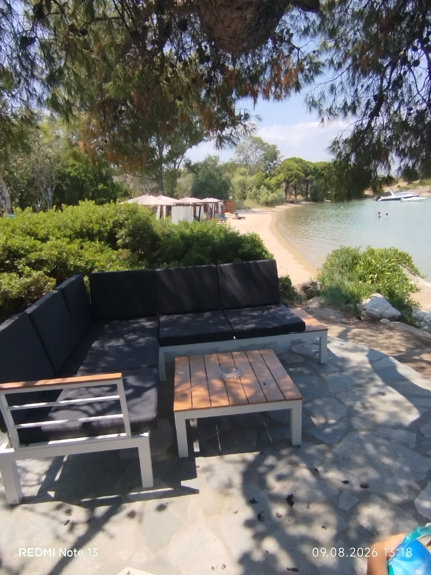 A scenic view of a beachside seating area at Sithonia Municipal Unit, Greece, is depicted. In the foreground, a modern outdoor sectional sofa with dark cushions sits on a stone-tiled patio. A matching wooden coffee table with slatted planks is placed in front of the sofa. To the left of the sofa, a single white-framed chair with wooden armrests is visible. The background reveals a sandy beach that curves gently into a clear blue body of water. Several white cabanas or tents are set up on the beach, and a line of lush green trees and bushes borders the sand. In the water, two boats are anchored, and two people are swimming. The sky above is a clear blue with scattered white clouds. The image captures a tranquil outdoor setting by the sea, with ample seating and a view of the water and beach.