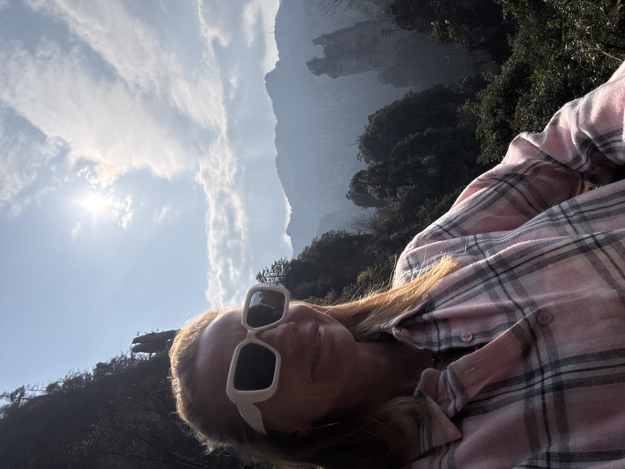 A mid-adult female is captured in a selfie outdoors in Wulingyuan, China. She is positioned in the foreground, with her head and upper torso visible. She has light-colored hair and wears large, rectangular, white-framed sunglasses with dark lenses, obscuring her eyes. A subtle smile is present on her face. Her attire includes a pink and gray plaid long-sleeved button-up shirt. A gold hoop earring is visible on her left ear. The background depicts the distinctive mountainous landscape of Wulingyuan, featuring numerous towering, tree-covered karst pillars and peaks under a bright blue sky with scattered white clouds. The sun is prominent in the upper-right quadrant, casting a strong glare and illuminating the scene. Distant mountain ranges appear hazy.