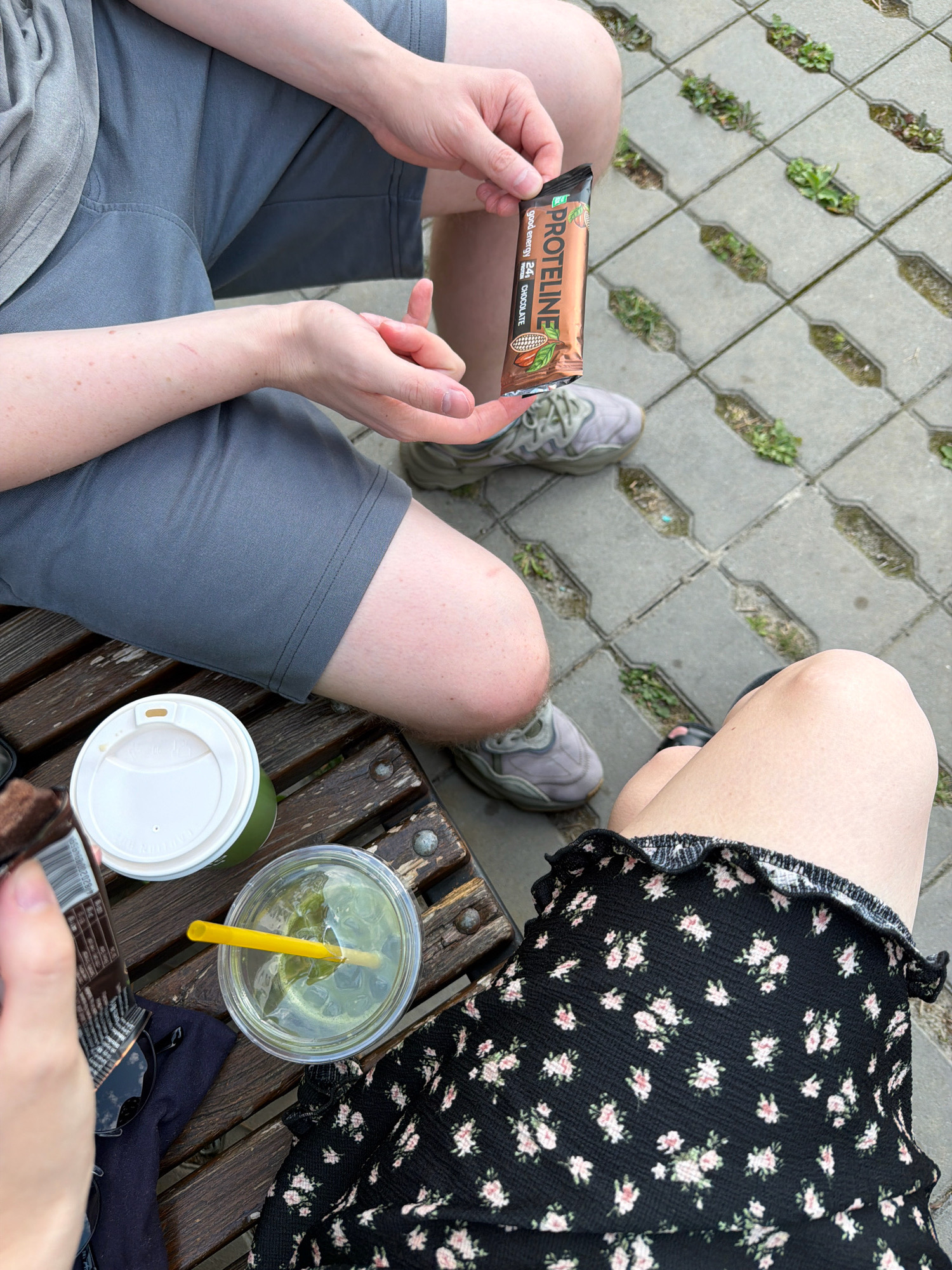 Two individuals are seated on a wooden bench. One person, wearing grey shorts and a grey t-shirt, is holding a chocolate-flavored protein bar. The other person is wearing a black floral skirt. On the bench between them are a white-lidded coffee cup and a clear plastic cup with a green beverage and a yellow straw. The background shows a paved area with rectangular stone tiles and patches of grass growing between them, typical of an urban park or public space in Kyiv, Ukraine. The overall scene suggests a casual outdoor break or meeting.