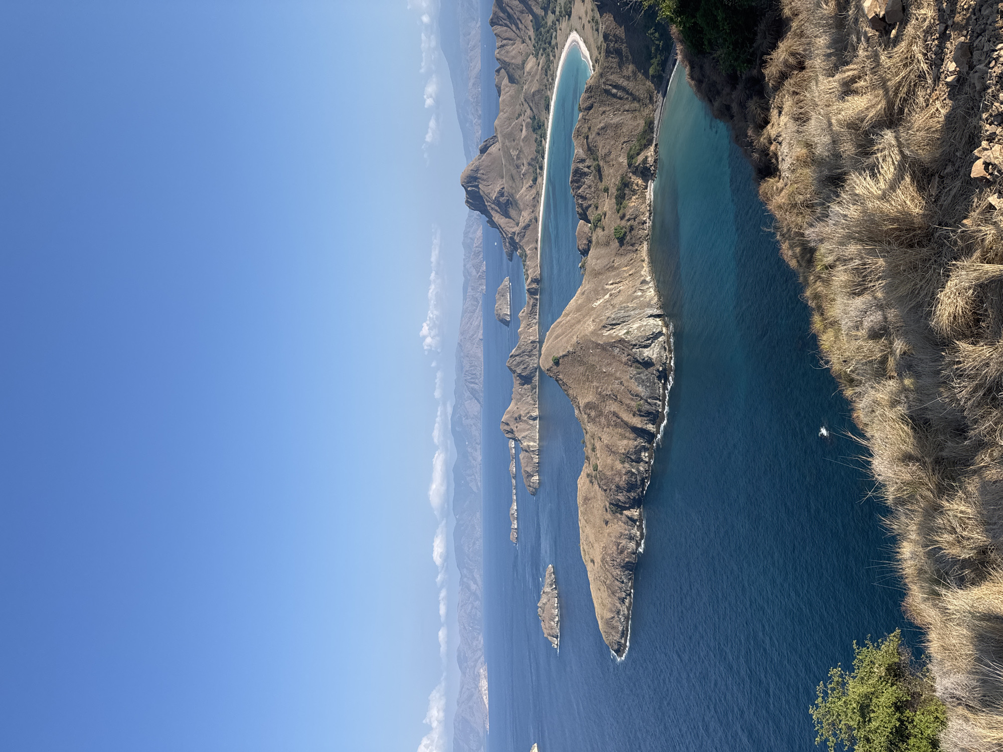 This is a high-angle, wide shot of a seascape with numerous islands under a clear blue sky. The foreground features dry, golden grass and rocks on a cliffside, overlooking the deep blue ocean. In the midground and background, several islands with sparse, dry vegetation and rocky shores are scattered across the water. The water ranges in color from deep blue to turquoise near the islands and beaches. In the far distance, a range of hazy, mountainous islands forms the horizon line, with a few wispy white clouds. The scene depicts a natural, uninhabited island landscape.