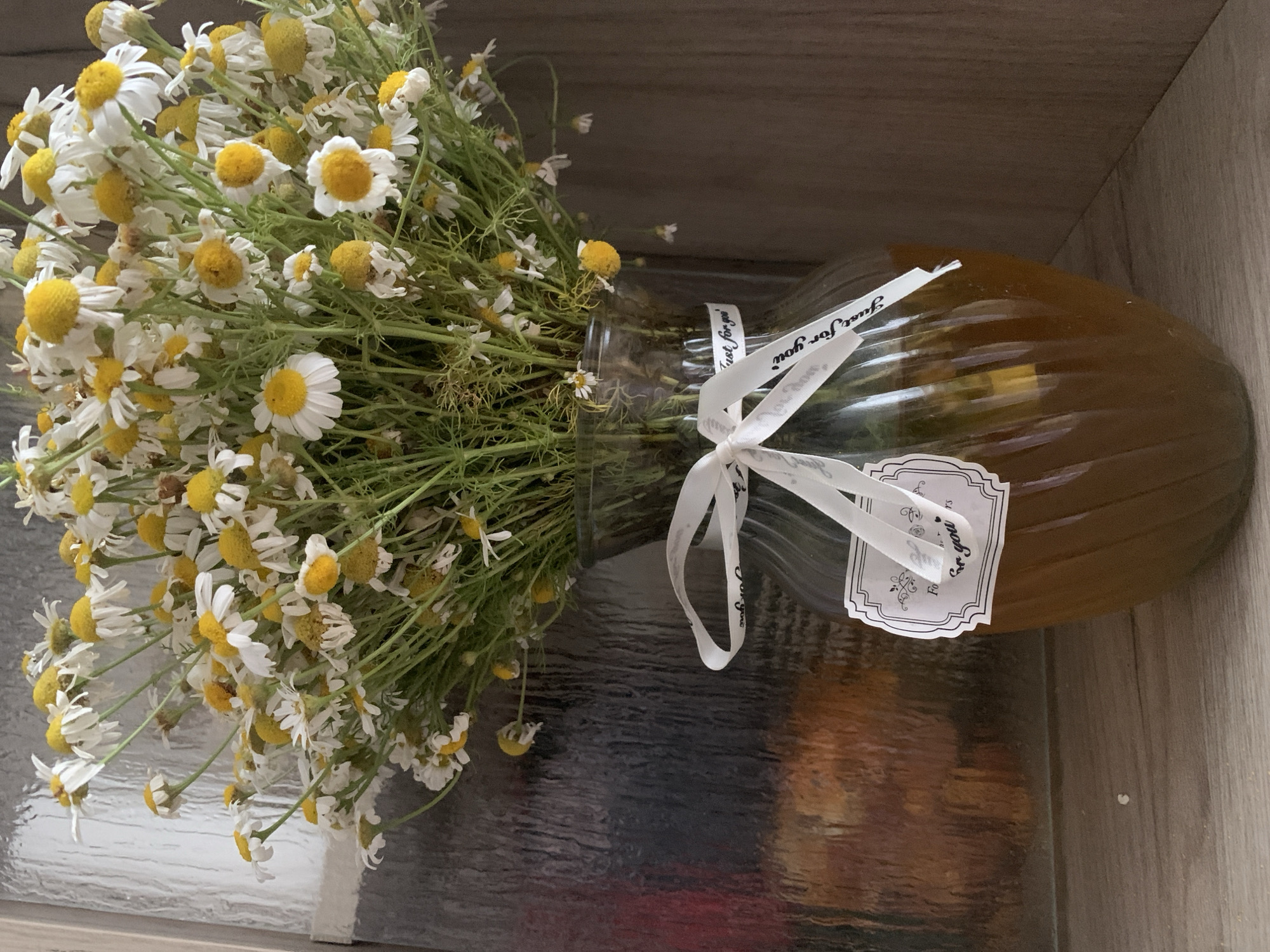 This media file features a bouquet of chamomile flowers in a glass vase. The vase is filled with a golden-brown liquid, possibly honey or tea, and is tied with a white ribbon. A decorative label on the vase reads "Just for you." The background is a wooden surface, suggesting an indoor setting. The lighting appears natural, casting soft shadows, and there are no indications of specific weather conditions or time of day. The overall scene evokes a sense of simple beauty and thoughtful gifting. The location context of Dondușeni, Moldova, is provided but not visually represented in the image.