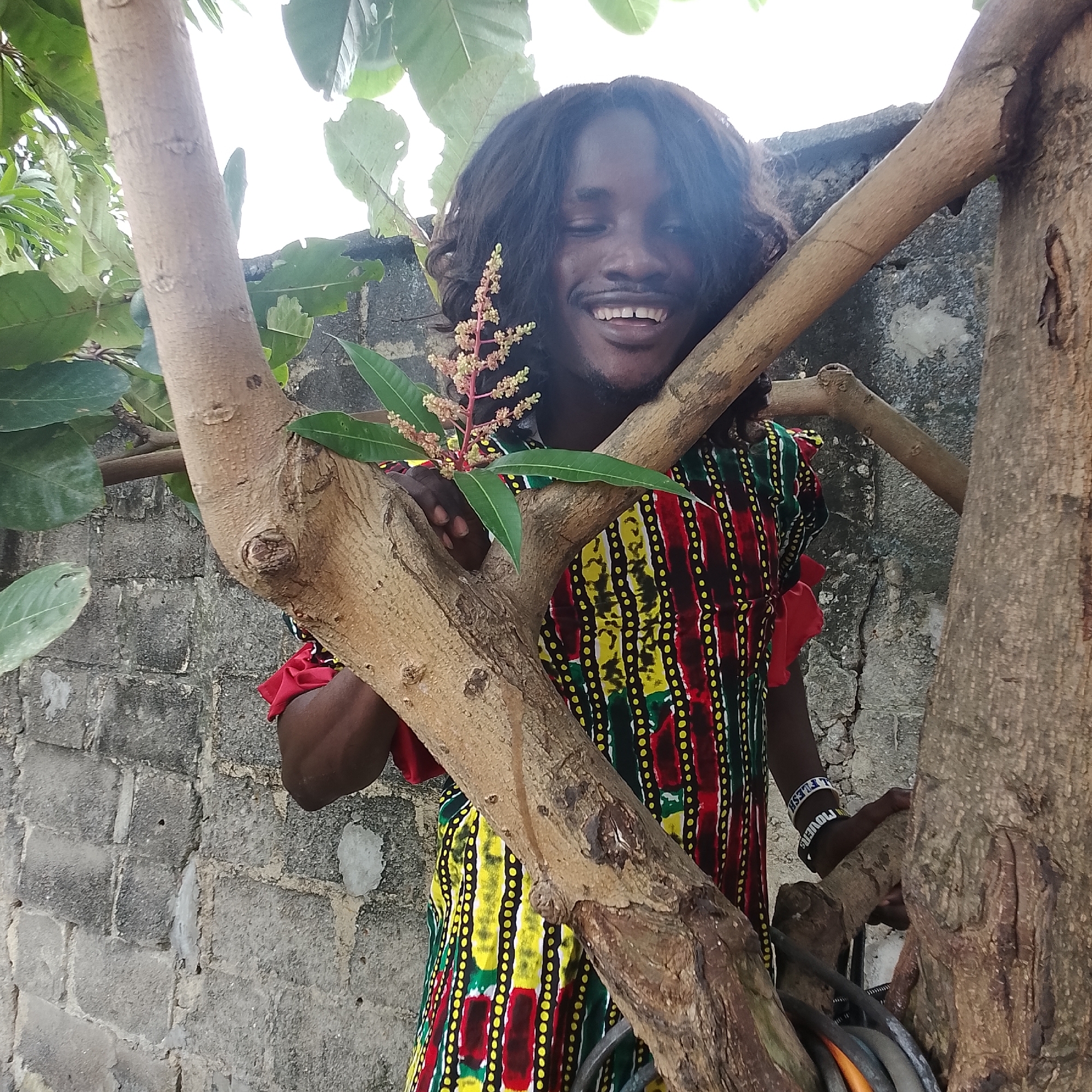 An outdoor, medium shot captures a Black male posing amongst tree branches in Ado, Nigeria. The man has dark, wavy hair and is smiling with his eyes closed, leaning his head against a tree trunk. He is wearing a colorful, patterned garment with vertical stripes of red, green, yellow, and black, accented with small yellow dots. His left hand is visible, holding a small branch with green leaves and a pinkish cluster of blossoms. On his right wrist, he wears two white beaded bracelets, one with black lettering that appears to say "FLESH" and another with the word "LOVERS". The background consists of a rough, gray cinder block wall and large green leaves from the surrounding trees. Two prominent tree branches are in the foreground, obscuring parts of the man and the wall. The lighting suggests it is daytime, with a bright sky visible through the foliage.