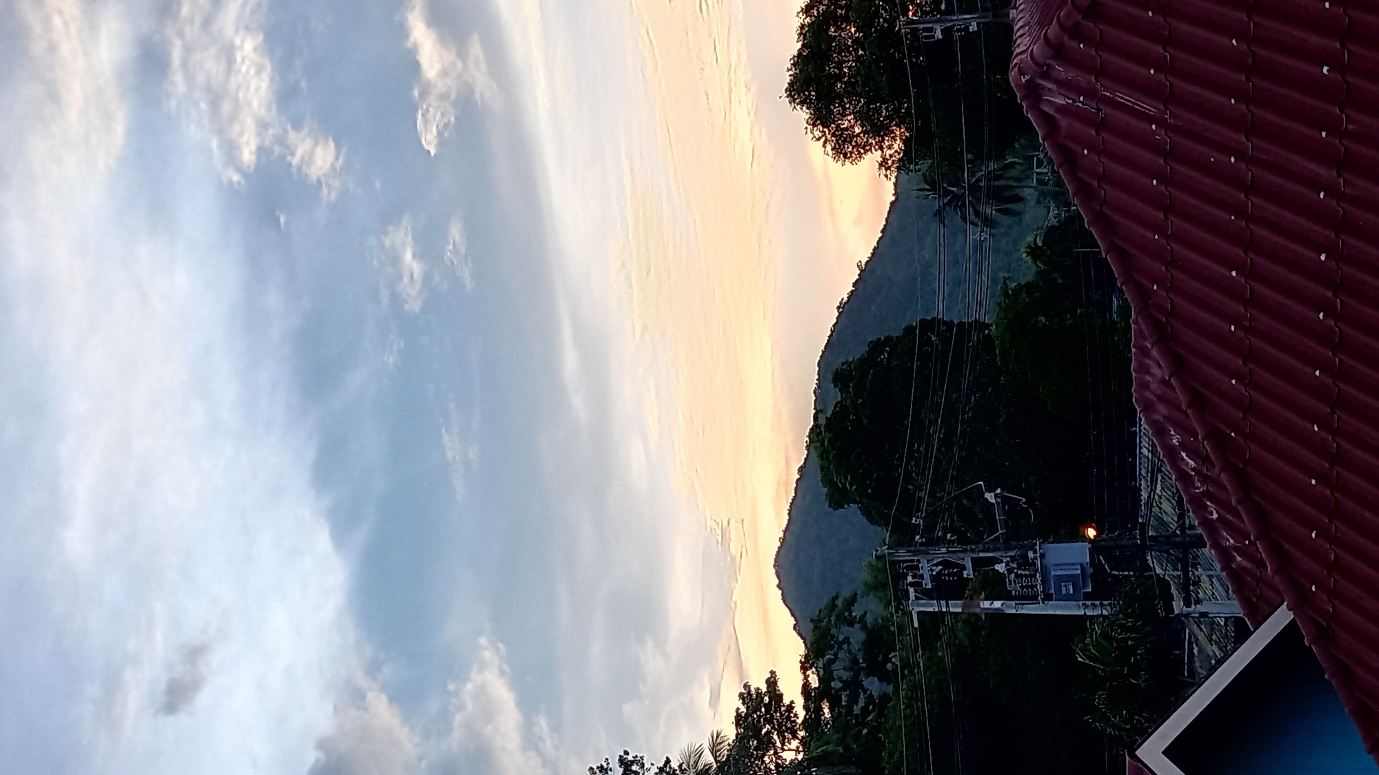 The image depicts a twilight scene in Ko Pha-ngan District, Thailand. In the foreground, a red tiled roof dominates the lower portion of the frame, with a glimpse of a blue-painted structure on the left. Behind the roof, a dense canopy of dark green trees lines the middle ground, interspersed with power lines and a utility pole featuring a transformer. Further in the background, a large, dark green mountain rises, its slopes covered in foliage. The sky above is a gradient of soft blues and pale oranges, with scattered wispy clouds, indicative of either sunrise or sunset. There are no people or animals visible in the image.
