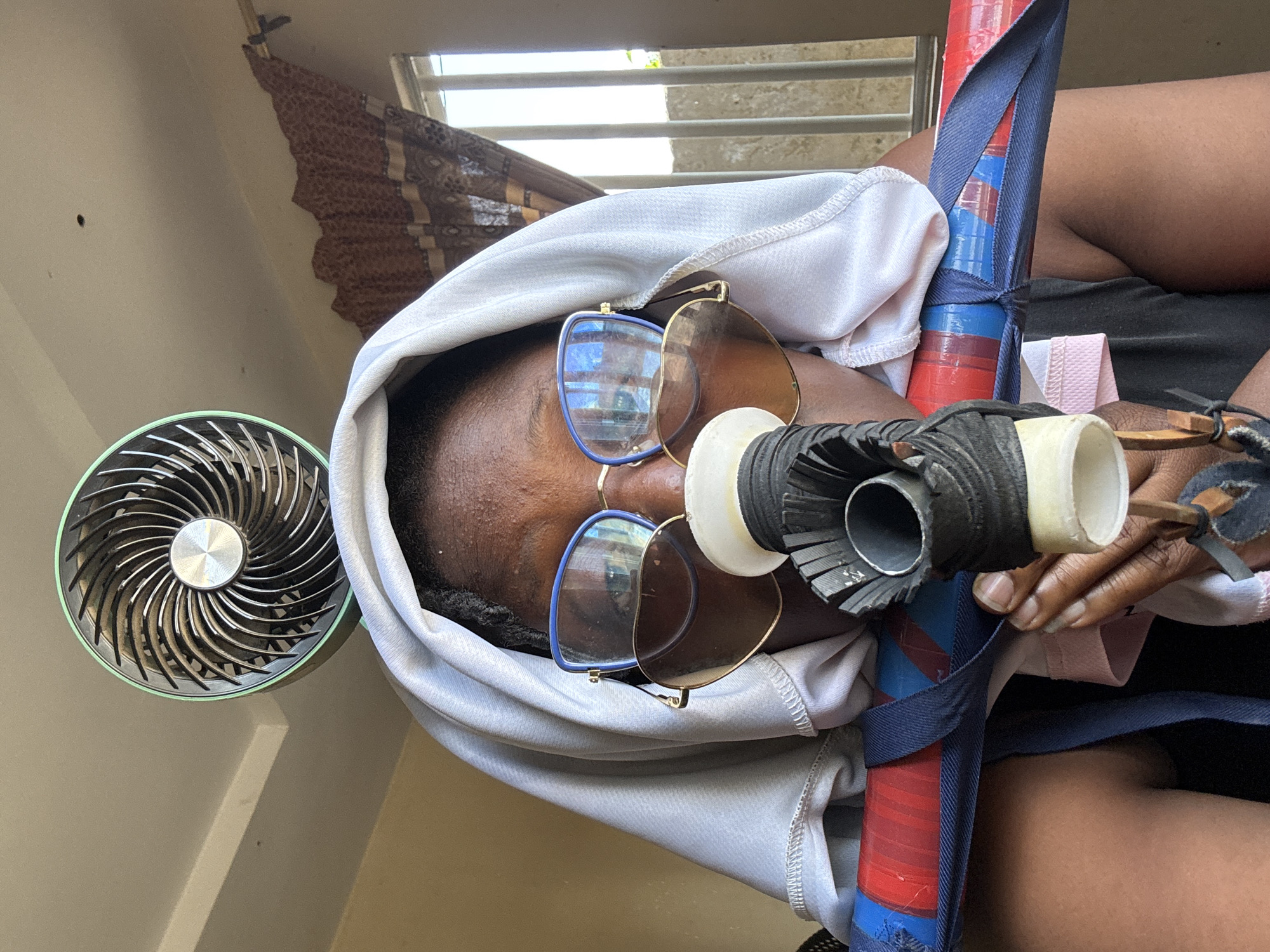 The image shows a close-up, front-facing view of a person wearing a white head covering, blue-rimmed glasses, and sunglasses. The person's face is partially obscured by a black and white apparatus attached to a red and blue tube, which is held across their chest. A small, circular fan with a green rim is positioned above and behind the person's head, against a beige ceiling. The background includes a window with bars and patterned curtains, suggesting an indoor setting. The scene is likely in Jawu, Nigeria, given the prompt. The overall impression is of an improvised setup, possibly for a performance, exercise, or artistic purpose. The lighting appears to be natural daylight coming from the window.