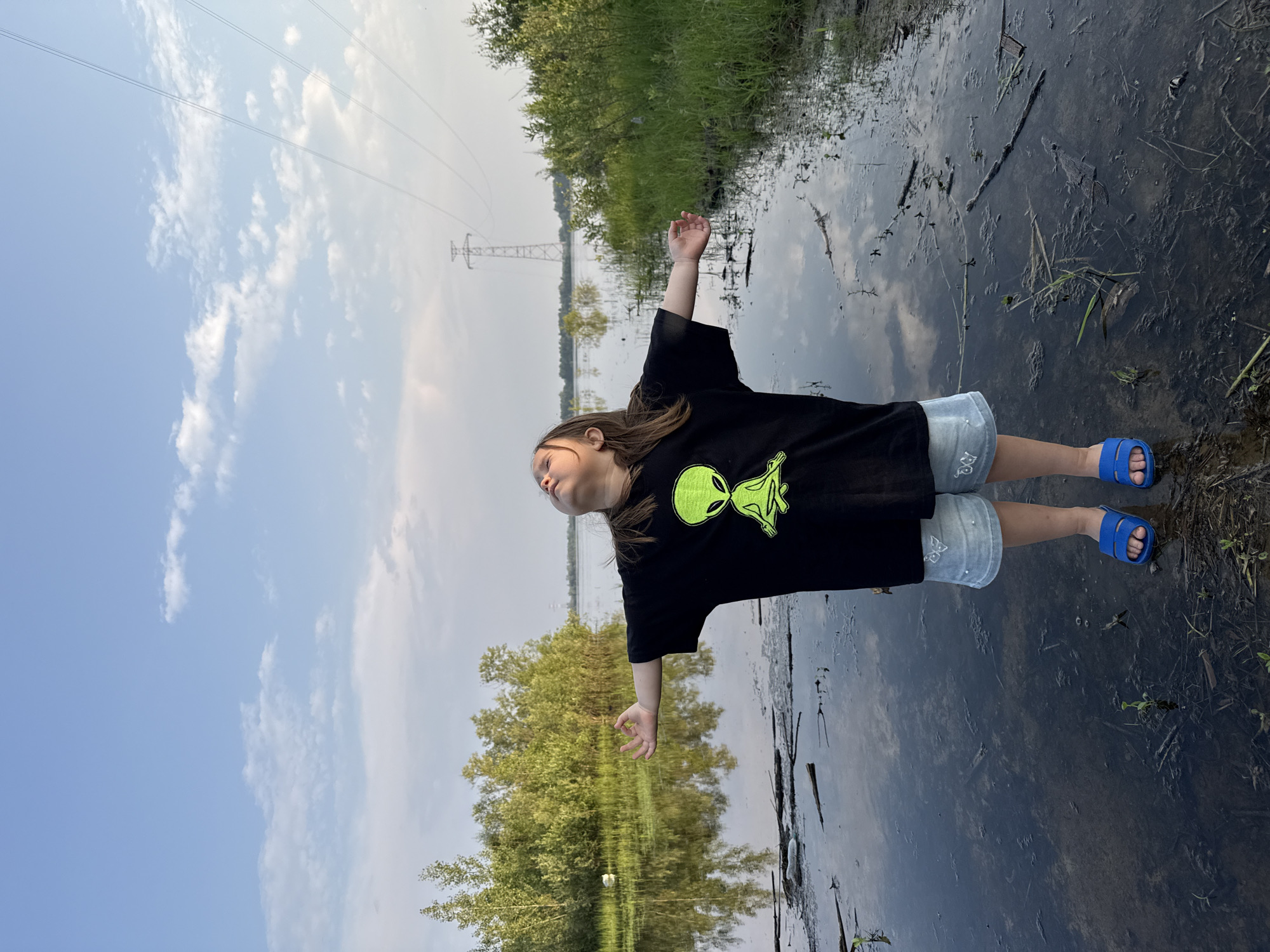 The image shows a young person, likely a child, lying down in shallow water with their arms outstretched. They are wearing a black t-shirt with a neon green alien graphic and light-colored shorts. Blue sandals are on their feet. The scene appears to be a natural outdoor setting, possibly a marshy area or a flooded field, with tall grass and trees visible in the background. The water reflects the sky and the surrounding vegetation. In the distance, a power line tower and wires are visible, indicating proximity to infrastructure. The sky is blue with scattered white clouds, suggesting a fair weather day. The overall mood seems peaceful and contemplative, with the child seemingly enjoying the natural environment. The orientation of the photo is unusual, as if taken from a low angle, or perhaps the camera was tilted.