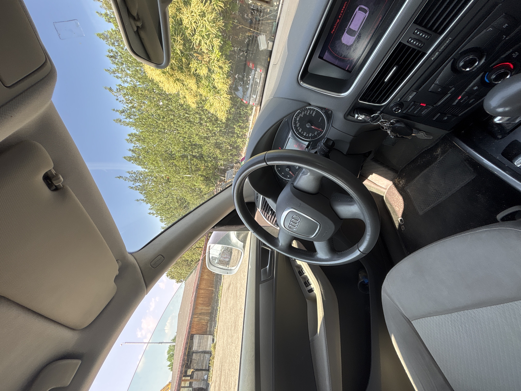 The image provides an interior view of an Audi car, specifically an Audi Q5 as indicated by the "Q5" text on the floor mat. The photo is taken from the passenger seat, looking across towards the driver's side and out the front windshield. The car appears to be stationary, parked outdoors on a bright and sunny day, with a clear blue sky and strong natural light casting shadows.
In the foreground, a gray and light gray two-tone fabric car seat is visible on the right. The steering wheel, featuring the prominent Audi rings logo, is centrally located, with car keys hanging from the ignition nearby. Behind the steering wheel, the instrument cluster displays a speedometer, tachometer, and other gauges.
The center console features an infotainment screen that shows a "Parking system" display. This screen illustrates a purple car outline with red and black proximity lines, suggesting the vehicle is either in the process of parking or has recently been parked. Below the screen are air conditioning vents, climate control dials with red and blue accents, and audio controls. The radio system is labeled "Audi concert," and the "RADIO" button is illuminated red. A gear shifter is partially visible on the right side of the console. The driver's side floor mat has some visible dirt.
Through the windshield, a vibrant green tree canopy dominates the exterior view. Beyond the trees, a dark-colored vehicle is discernible. On the far right, a wooden structure, possibly a fence or building, and a yellow construction vehicle (likely an excavator arm) are visible. The passenger-side sun visor is lowered. A small, square, clear object or reflection is present on the upper part of the windshield. The A-pillar trim bears the text "AIRBAG." No people are visible in the scene.