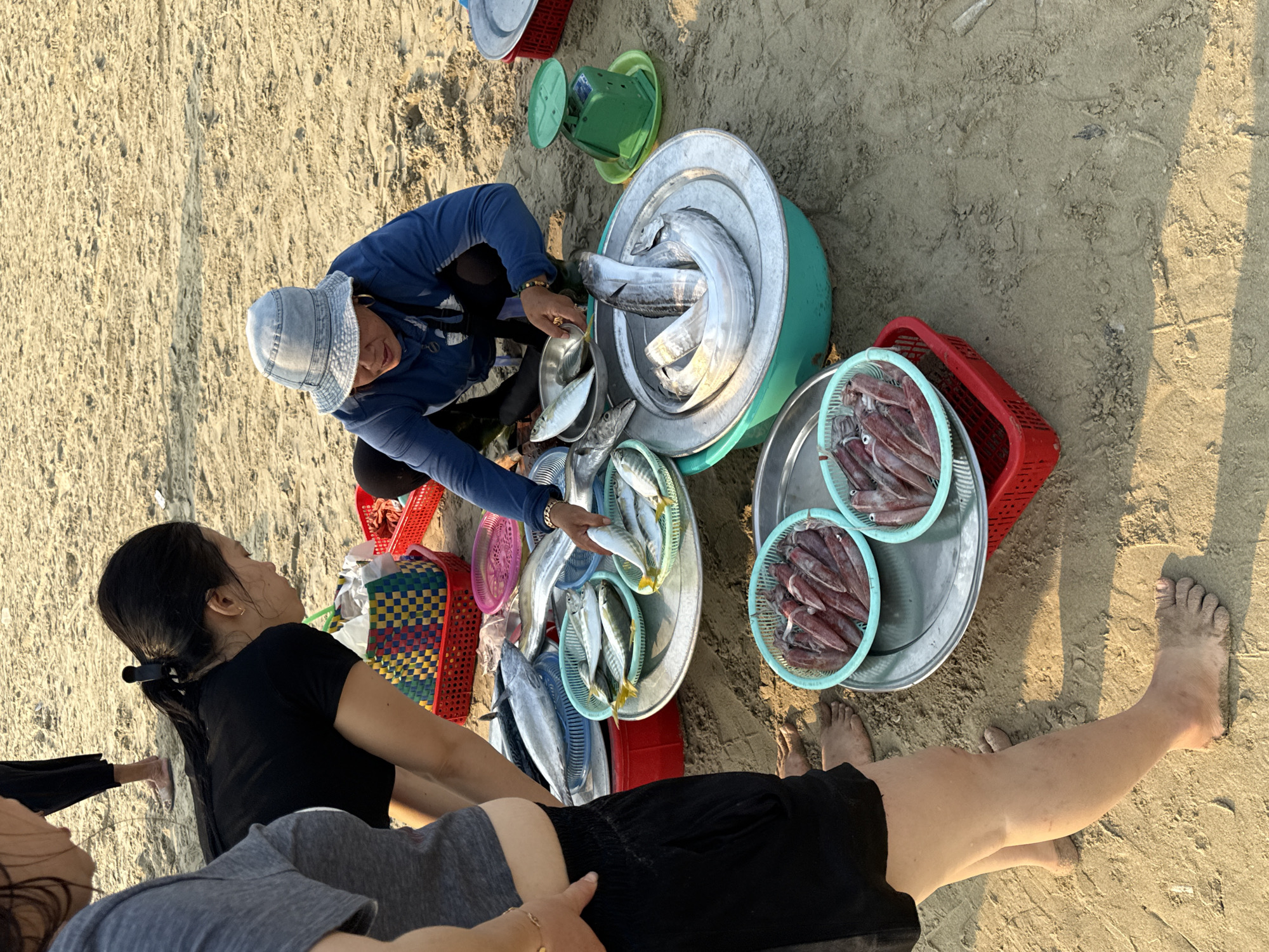 The scene is outdoors on a sandy beach in Đà Nẵng, Vietnam. A woman with a denim hat and blue jacket is selling fish and squid. She is squatting and handling fish, placing them in a metal dish. A customer is leaning forward, looking at the seafood. Baskets of fresh seafood, including fish and squid in light blue plastic baskets, are arranged around her. A green scale is visible near the fish. The sand has a light beige tone. A person in black shorts lies on the sand with visible feet and legs.