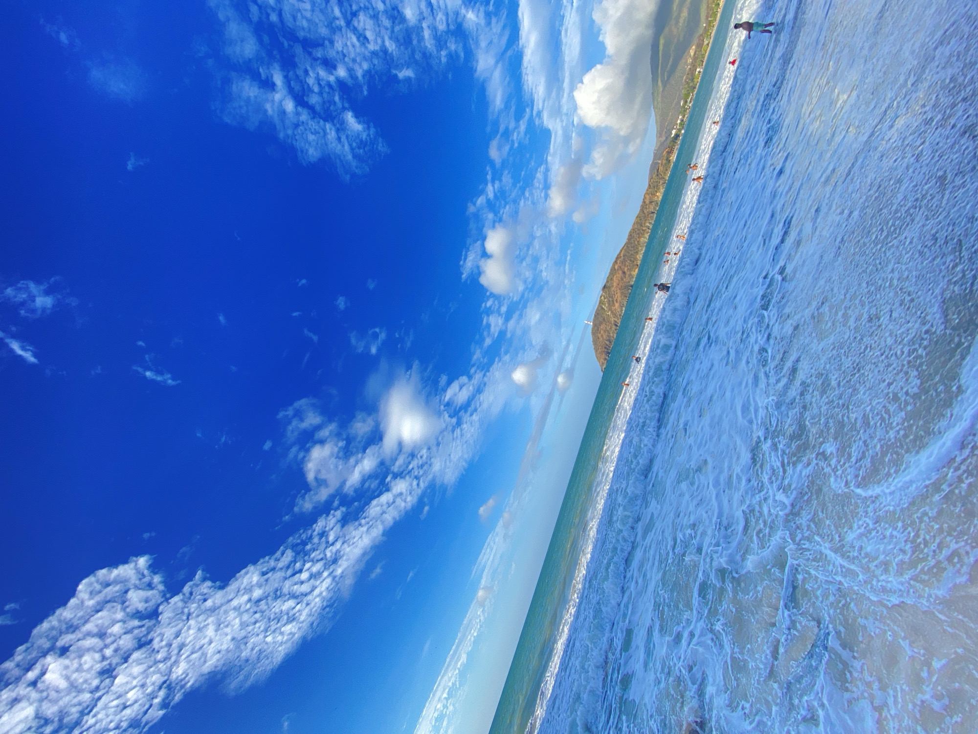 The image captures a coastal scene in a bright, daytime setting. A vibrant blue sky with scattered white clouds occupies a significant portion of the view. The horizon line separates the sky from the ocean, which is actively churning with foamy waves washing onto the sandy beach. Several people are seen along the shoreline, some seemingly wading in the water. In the background, a mountainous coastal landscape extends along the coast. The photograph seems to have been taken at a slightly tilted angle, giving it a dynamic perspective. The location in the city of Pedro González, Venezuela cannot be confirmed using visual data alone.