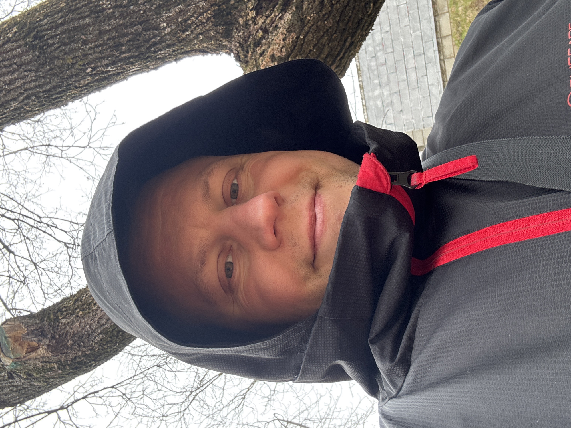 A man wearing a black jacket with red accents is facing the camera. He is hooded and appears to be outdoors, as a tree trunk and branches are visible in the background. The surroundings suggest an urban area, with what looks like a brick or stone paved surface partially visible. There is no specific evidence to confirm the location is Minsk, Belarus.