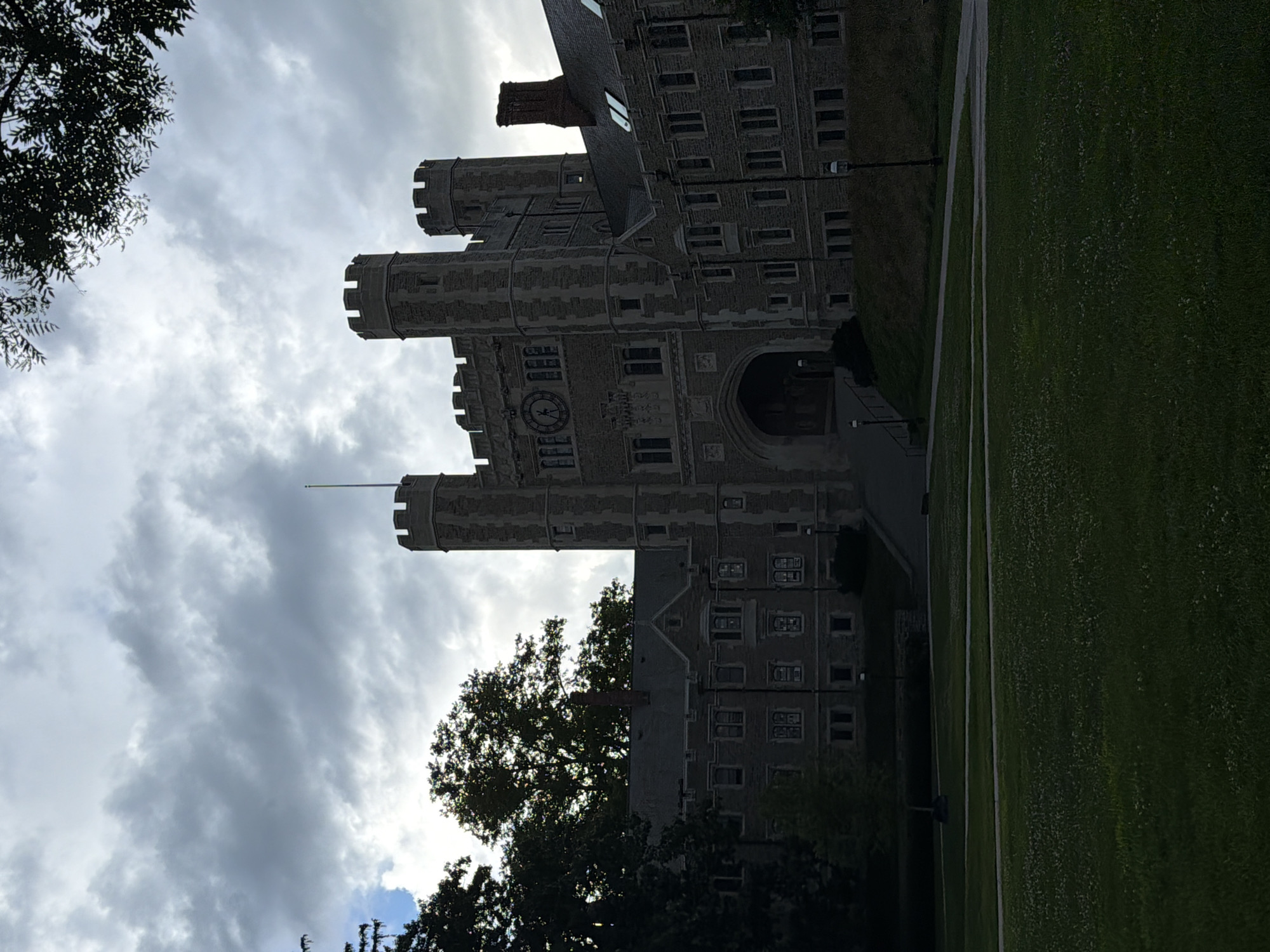 An overcast day looms over the gothic architecture of Princeton University's Nassau Hall in Princeton, New Jersey, USA. The imposing stone building, with its distinctive clock tower and crenellated turrets, is silhouetted against a cloudy sky. The foreground is dominated by a lush, green lawn dotted with tiny white flowers, leading up to a paved walkway that curves towards the building's entrance. Trees frame the scene, their dark foliage contrasting with the lighter stone of the hall and the diffused light filtering through the clouds. The atmosphere is serene and somewhat somber, reflecting the muted light and the grand, timeless nature of the historic university.