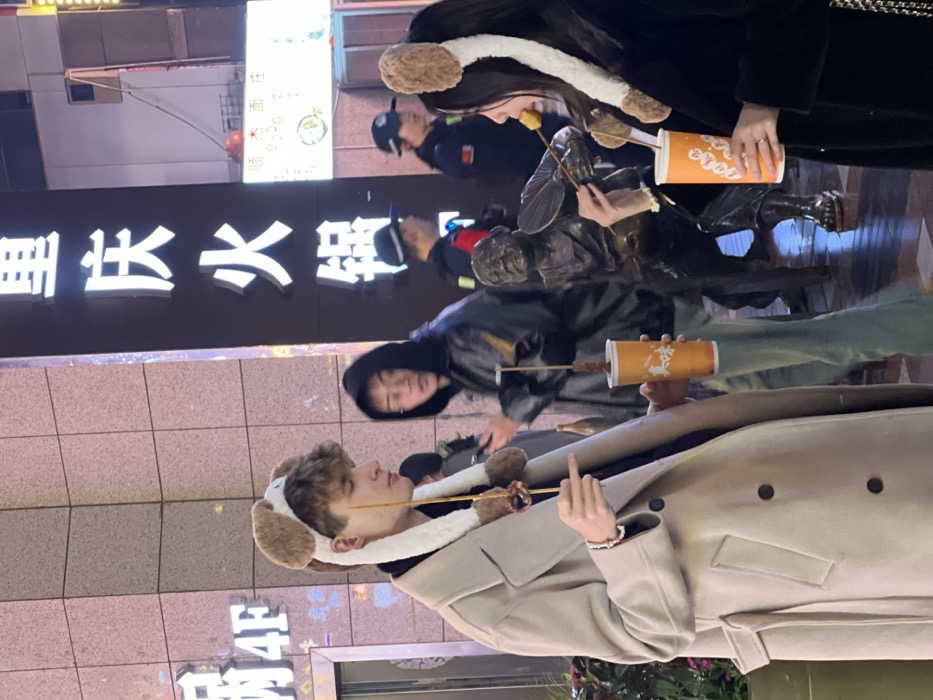 The image displays an urban street scene at night in Yuzhong District, Chongqing, China, under artificial illumination. In the foreground, a young male with light brown curly hair wears a light beige double-breasted overcoat and a plush light brown and white dog-ear headband with long flaps. He holds a wooden skewer with two dark food items, pointing downwards, in his right hand. His left hand holds an orange cup containing another skewered dark food item. He is looking towards his right. Adjacent to him on the right, a young female with dark hair wears a dark coat and a similar dog-ear headband. She holds an orange cup with a skewer containing a light yellow food item in her left hand and is actively consuming it. She looks towards her left.
Between these two individuals, slightly behind, stands a bronze statue of an elderly man, depicted holding a fan in his left hand and a skewer with food items in his right hand. In the middle-ground, a male of East Asian descent, wearing a dark hooded jacket and glasses, looks towards the foreground male with an open-mouthed expression. He also appears to be holding an orange cup with a skewer.
The background features building facades. On the right, a prominent vertical sign displays illuminated white Chinese characters "重庆火锅" (Chongqing Hotpot). On the left, another sign partially visible indicates "4F". Further in the background, several blurred pedestrians are visible, including two individuals in dark uniforms with caps and reflective armbands, identifiable as security personnel or police officers. The ground is paved, and the area suggests a commercial district.