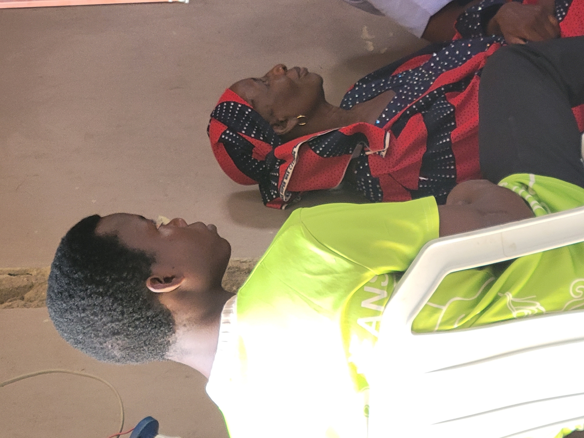 Two individuals are visible in this image. The first individual, an older woman, is lying on the floor, dressed in a patterned red and black headwrap and a matching top, with black leggings. She is lying on her back. The second individual, a younger man, is sitting in a white plastic chair and wearing a bright green t-shirt. The location appears to be indoors, likely in Madalla, Nigeria. The floor has a plain, mottled surface. Additional details include the presence of a wire and a small blue and red object on the floor near the young man.