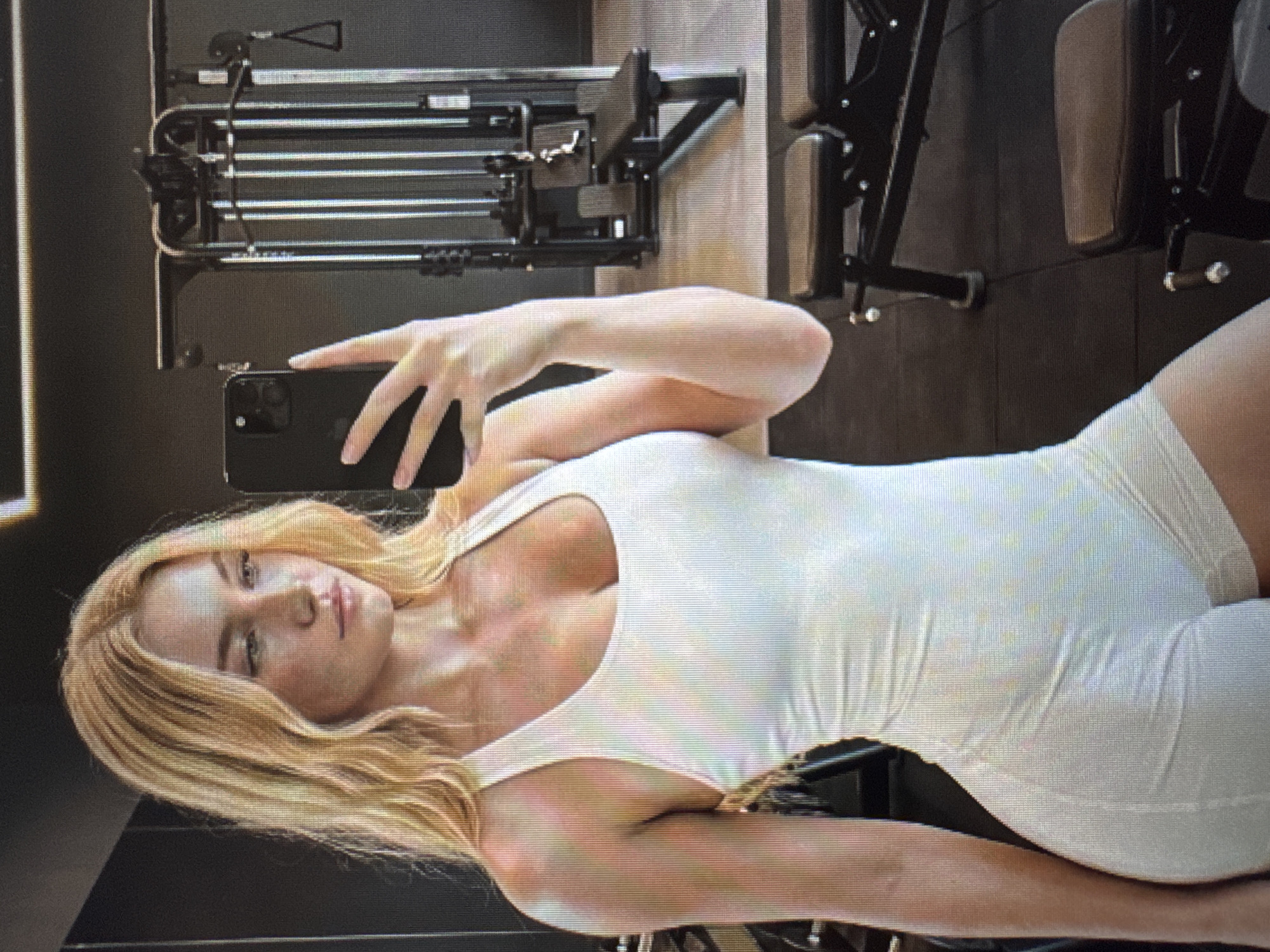This rotated image captures a young woman taking a mirror selfie in what appears to be a gym or fitness center. The woman, with long, wavy blonde hair, stands with a confident, neutral expression, looking directly at her reflection. She is wearing a form-fitting, sleeveless white bodysuit or jumpsuit. Her right arm is raised, holding a black smartphone, possibly an iPhone due to the visible camera module, to take the picture.
In the background, various pieces of gym equipment are visible, including multiple weight machines with cables, pulleys, and padded components, suggesting a dedicated strength training area. The equipment is dark-colored, likely black or grey metal. The setting is indoors, with ambient lighting illuminating the scene. No discernible text is present on the woman's clothing or the gym equipment. The image quality shows some pixelation, particularly noticeable on closer crops.