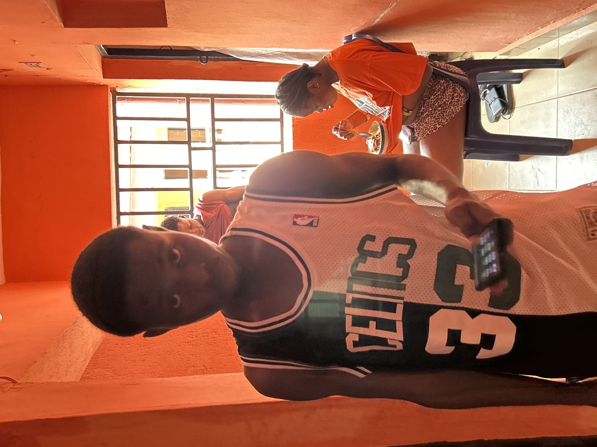 The image shows three people in an orange-painted room in Ihiagwa, Nigeria. One person is wearing a Boston Celtics jersey and holding a cell phone, looking directly at the camera. Another person sits at a table eating from a bowl. A third person is visible in the background. A window with bars is present. A bag is hung on the back of the chair.
