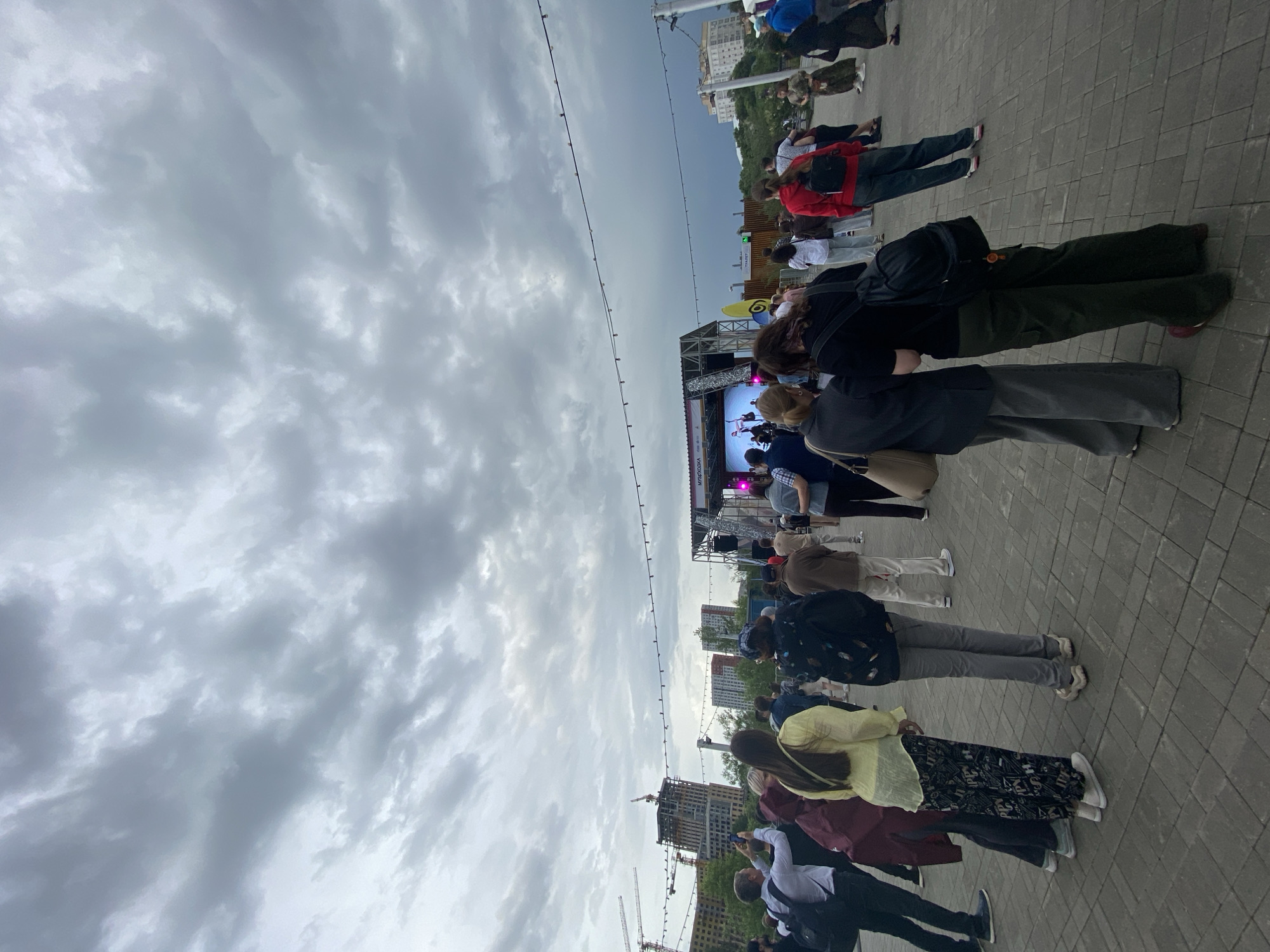 The image depicts an outdoor public gathering in an urban setting, likely Tyumen, Russia, viewed from behind a crowd looking towards a stage. The sky is heavily overcast with a thick layer of grey clouds.
Numerous people are visible, standing on a paved surface made of grey and tan interlocking blocks. They are casually dressed and appear to be attentive to a stage located in the middle distance. The stage features a large screen displaying what appears to be a performance or presentation, with images of people on it. Cyrillic text, notably "МУЗЫКА" (Music), is visible on the lower part of the screen or stage structure, indicating a musical event.
Above the crowd, a string of lights or cables stretches horizontally across the scene. In the background, behind the stage, various city buildings are visible, including some under construction with cranes, confirming an urban environment. The time of day is not clearly indicated but the diffused light suggests a cloudy day, possibly late afternoon or early evening. The overall atmosphere is that of a public outdoor festival or concert.