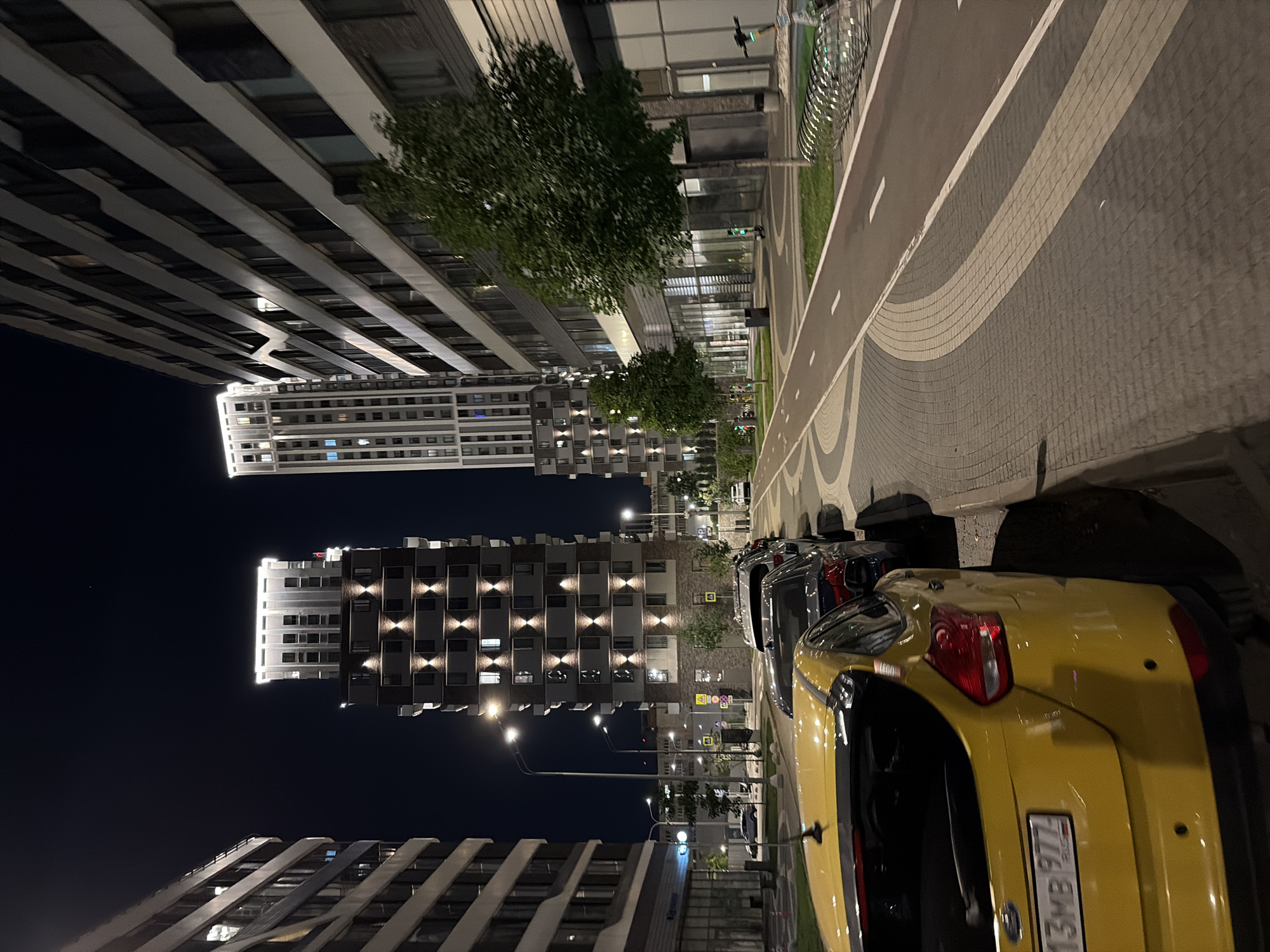 This image shows a nighttime street scene in Moscow, Russia. The foreground features the rear of a bright yellow car parked along a paved sidewalk, with the license plate reading "13MB 977". Several other cars are parked in a row behind it. The street itself has a designated bike lane with white markings. Flanking the street are modern apartment buildings. One tall building on the left is mostly dark with some illuminated windows, while the building in the center is a taller, slender structure with prominent upward-pointing lights accentuating its facade. To the right, a long, multi-story building with many windows dominates the frame. Trees are planted along the sidewalk, and streetlights illuminate the scene. In the background, more buildings and streetlights are visible, suggesting a developed urban environment. There are no people visible in the image.