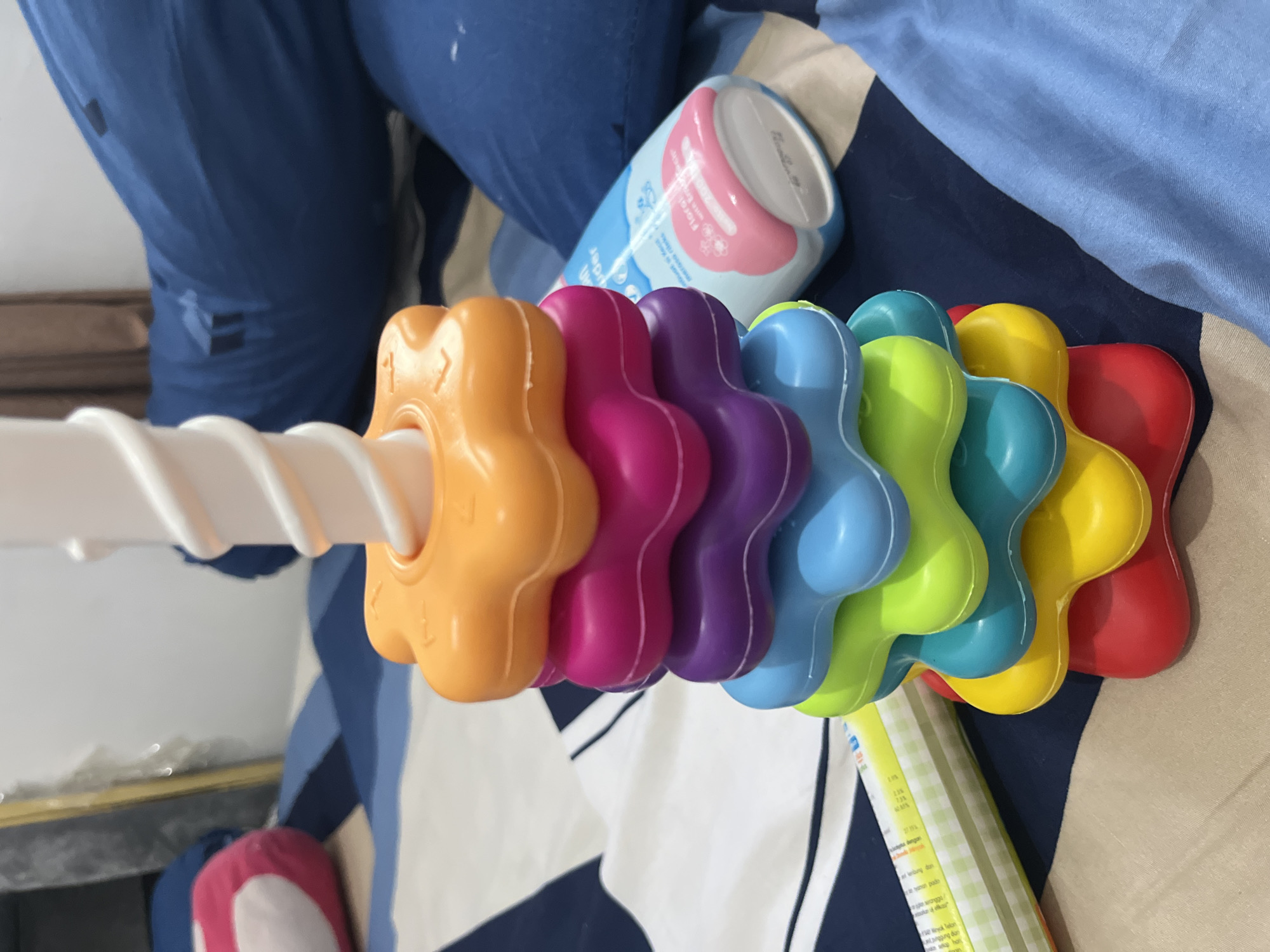 The image displays a close-up, high-angle view of baby-related items resting on a patterned fabric surface, likely a bed, in an indoor setting in Lhokseumawe, Indonesia.
The central subject is a colorful plastic baby toy, appearing to be a rattle or stacking toy. It features several flower-shaped pieces in a sequence of vibrant colors: orange, magenta, purple, light blue, lime green, yellow, and red, all threaded along a white spiral rod.
In the background, a person's leg or body, covered in blue fabric with a dark blue design, is partially visible. To the upper right of the toy, a light blue bottle with a pink and white cap is present. Visible text on the bottle includes "WAMS", "Puder", "Floral", and parts of "meroso rileks", indicating it is likely a baby powder or lotion product. Below the toy, a light yellow rectangular object with green stripes, possibly a book or product packaging, is visible. Text on this object includes "ryok lelon", "nanggung dan", "te tyanon pado", "23%", "733", and fragments such as "dengan lembut dan". The fabric beneath the items features a geometric pattern of dark blue, light blue, and beige sections.
The scene depicts a static arrangement of items, suggesting a domestic and nurturing environment associated with baby care. There is no specific activity occurring.