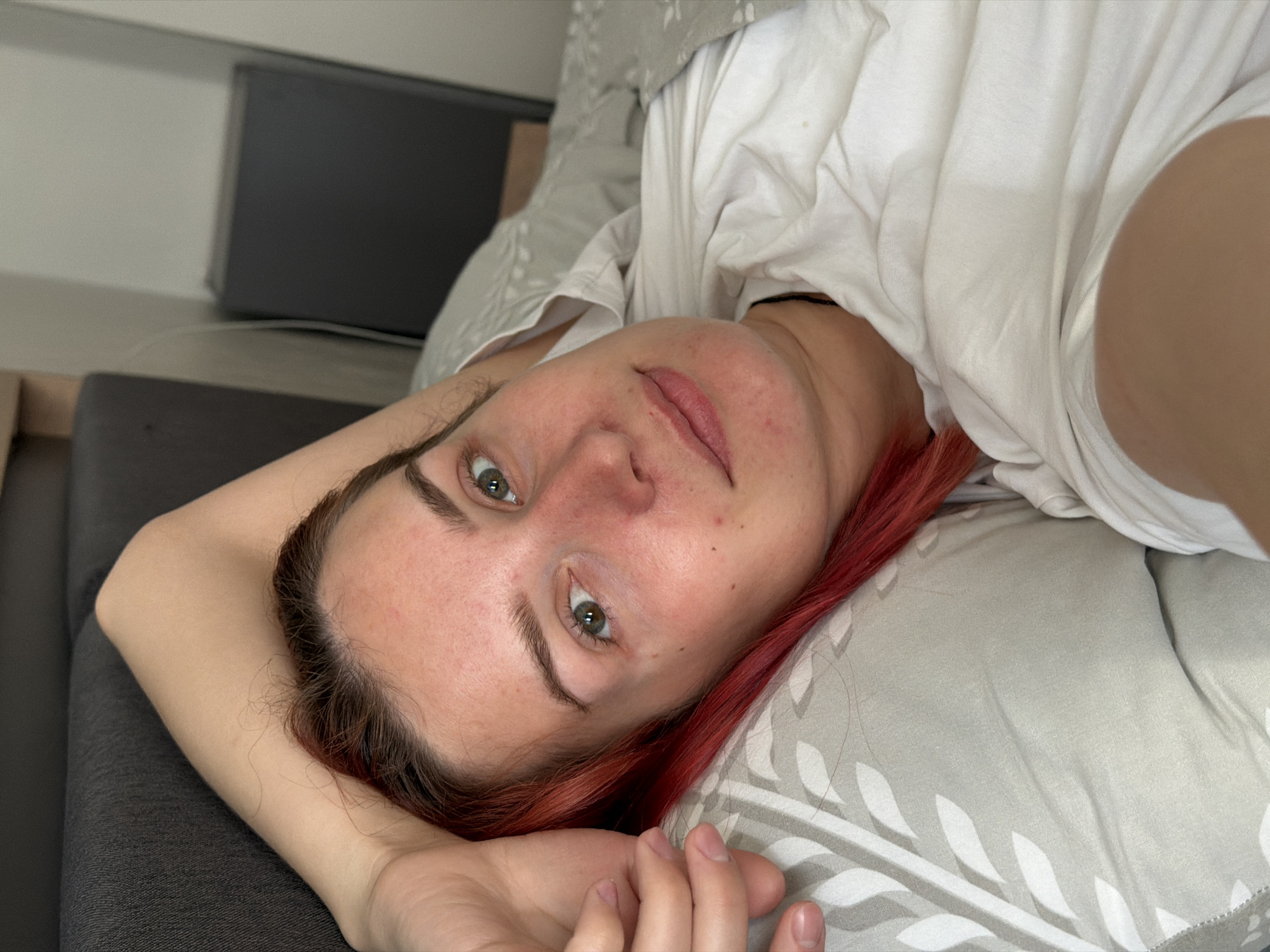 A woman with red hair lies on a bed in an interior setting. She wears a white t-shirt and is positioned on a gray pillow with a leaf pattern. The environment includes a gray couch and a gray cabinet in the background. The location is unspecified, and there is no indication it is in Naberezhnye Chelny, Russia.