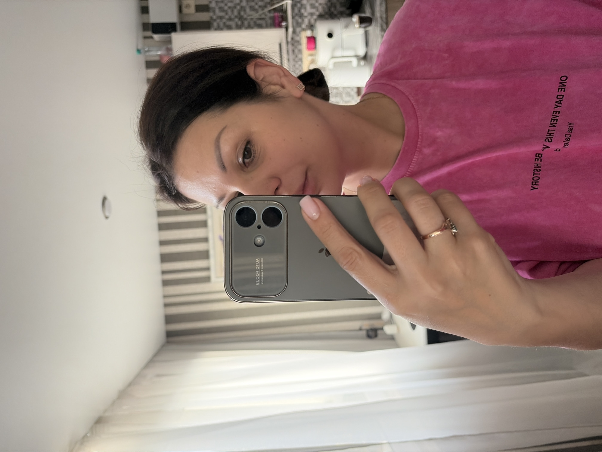 A medium shot captures a woman taking a selfie in a brightly lit room. She is positioned in the center-right of the frame, holding a silver smartphone in her left hand to photograph her reflection in a mirror. Her head is turned towards the camera, with her dark hair pulled back. She wears a vibrant pink t-shirt with text printed upside down and backward: "ONE DAY EVEN THIS V." and "BE HISTORY." A delicate gold ring with a small stone is visible on her left ring finger.
The background shows a window with sheer white curtains on the left and a striped wallpaper pattern of light gray and white on the right. In the background, a kitchen counter is partially visible with appliances including a white food processor or air fryer, and bottles.
The location is inferred to be Sochi, Russia, based on the provided context. The scene appears to be an indoor domestic setting, likely a home.