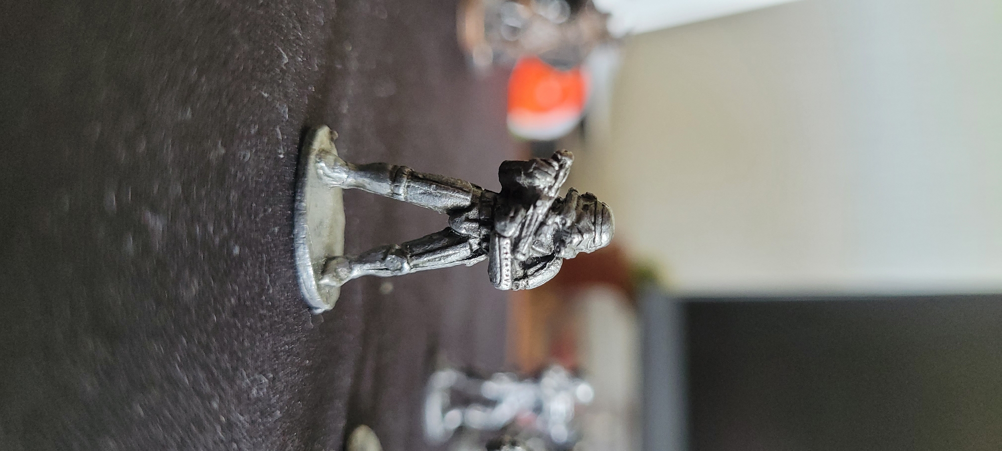 A close-up, shallow depth-of-field shot captures a miniature metal figure of a Stormtrooper from Star Wars. The figure is standing on a dark, textured surface, possibly a table or mat, and is posed with a rifle. The background is blurred, showing hints of other figures and a light-colored wall or object. The lighting is natural and casts soft shadows. The image was taken indoors, likely in a residential setting, with no indication of the city of Heusenstamm, Germany.