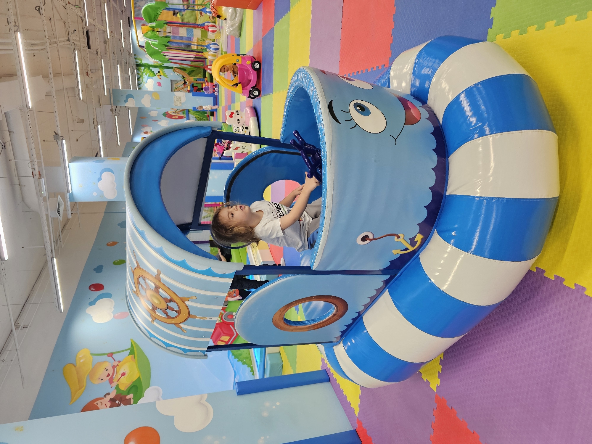 A child is playing inside a blue inflatable boat in a children's playroom. The boat is decorated with a smiling face and has blue and white striped inflatable bumpers. A steering wheel is inside the boat. The playroom has colorful floor mats and murals on the walls with cartoon-like characters. Other play structures can be seen in the background. The location is in Khimki, Russia.