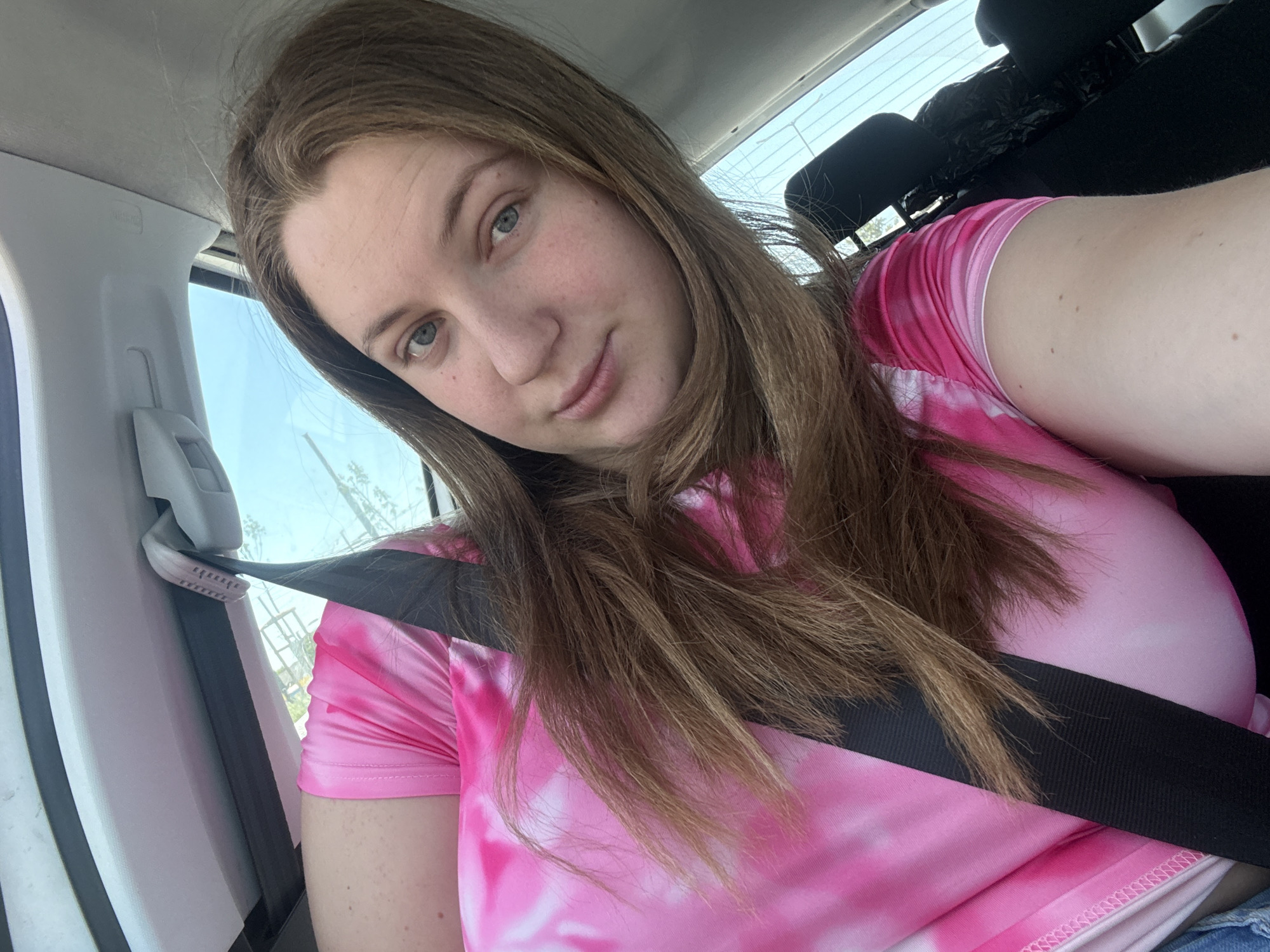 The image shows a person in the driver's seat of a vehicle. They are wearing a pink tie-dye shirt and have a seatbelt fastened across their chest. The vehicle is likely stationary, as the person is taking a selfie. The location in the city of Unknown, Unknown cannot be determined from the image.