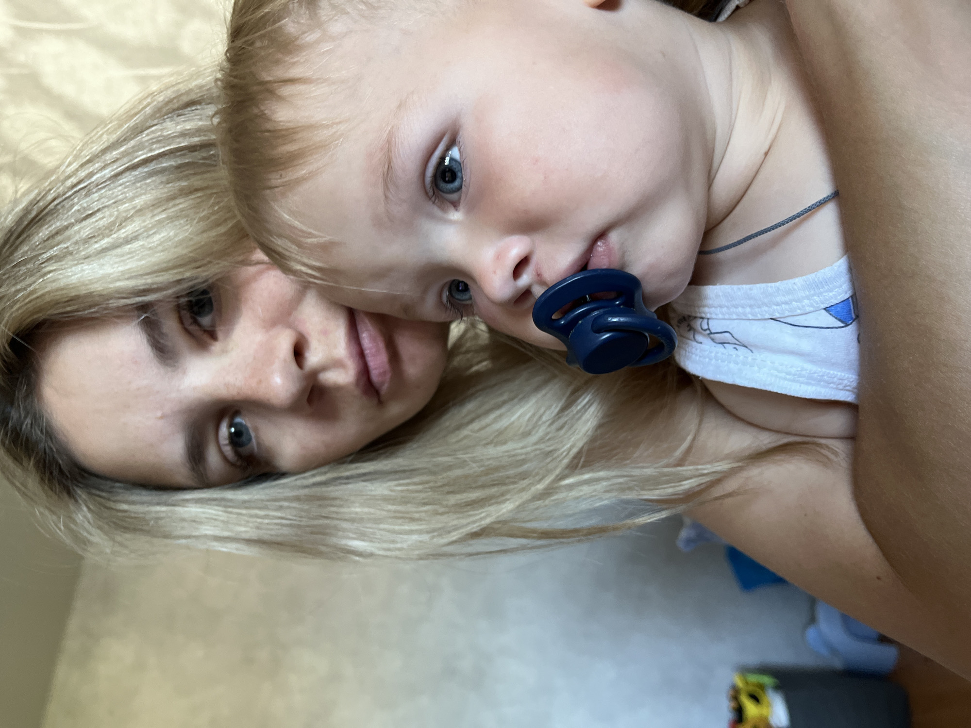 A woman with blonde hair and blue eyes is holding a baby with blonde hair and blue eyes. The baby has a pacifier in its mouth. They are both looking towards the camera. The background is a wall with a light-colored texture. The image appears to be taken indoors. There is a faint shadow on the wall. There are no discernible landmarks or elements that would specifically place this image in Chaikovsky, Russia, or any other specific city. The scene depicts a maternal moment, with the woman holding the baby close. The lighting is natural and appears to be from a window or overhead light.