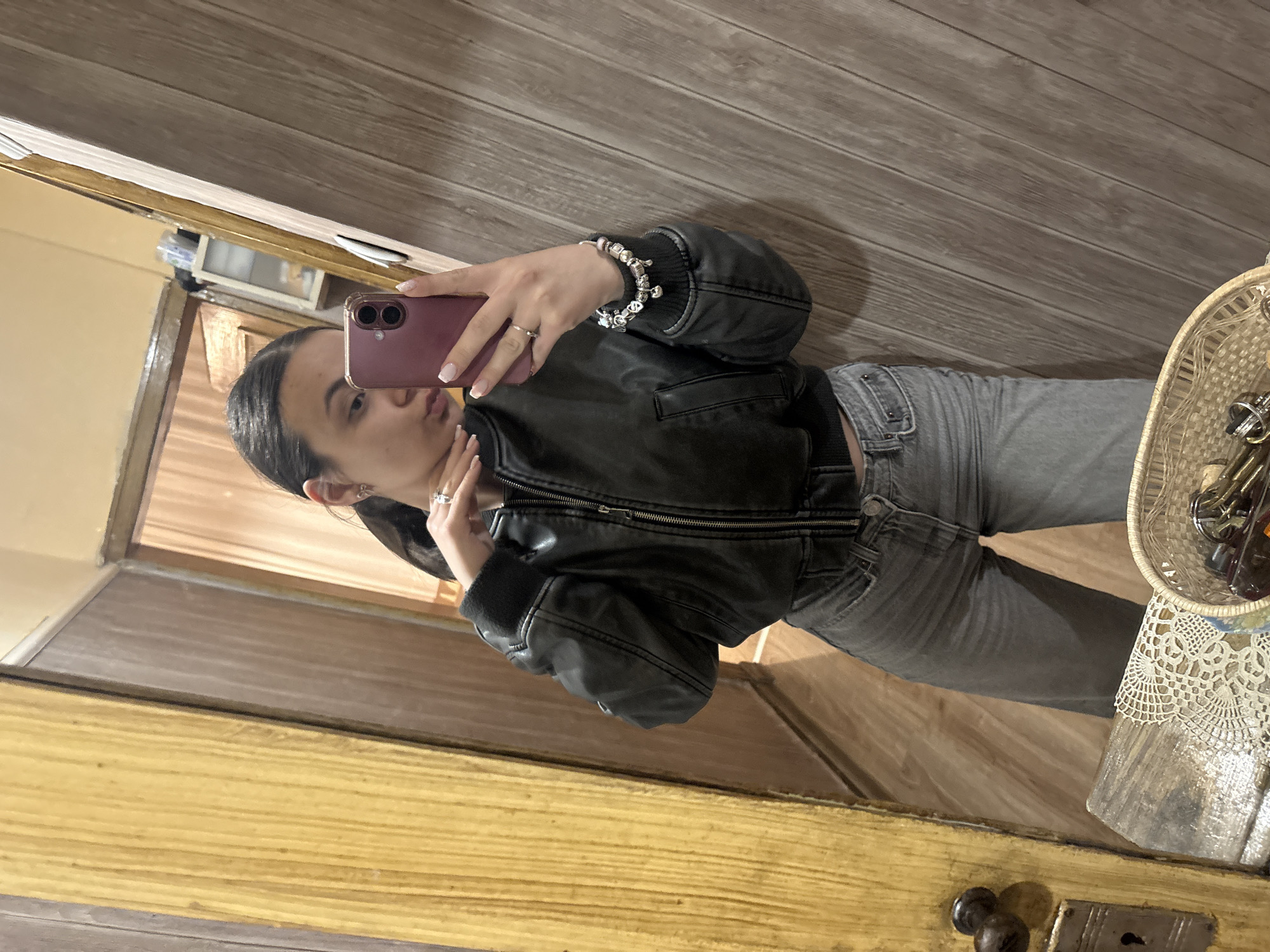 A woman is taking a selfie in front of a mirror. She is wearing a black leather jacket, gray jeans, rings, and a bracelet. The background includes a wooden wall, a partially visible open doorway, and a wooden chest of drawers. Inside a wicker basket, atop the chest of drawers, are keys. There are no details to indicate this photo was taken in Radomir, Bulgaria.