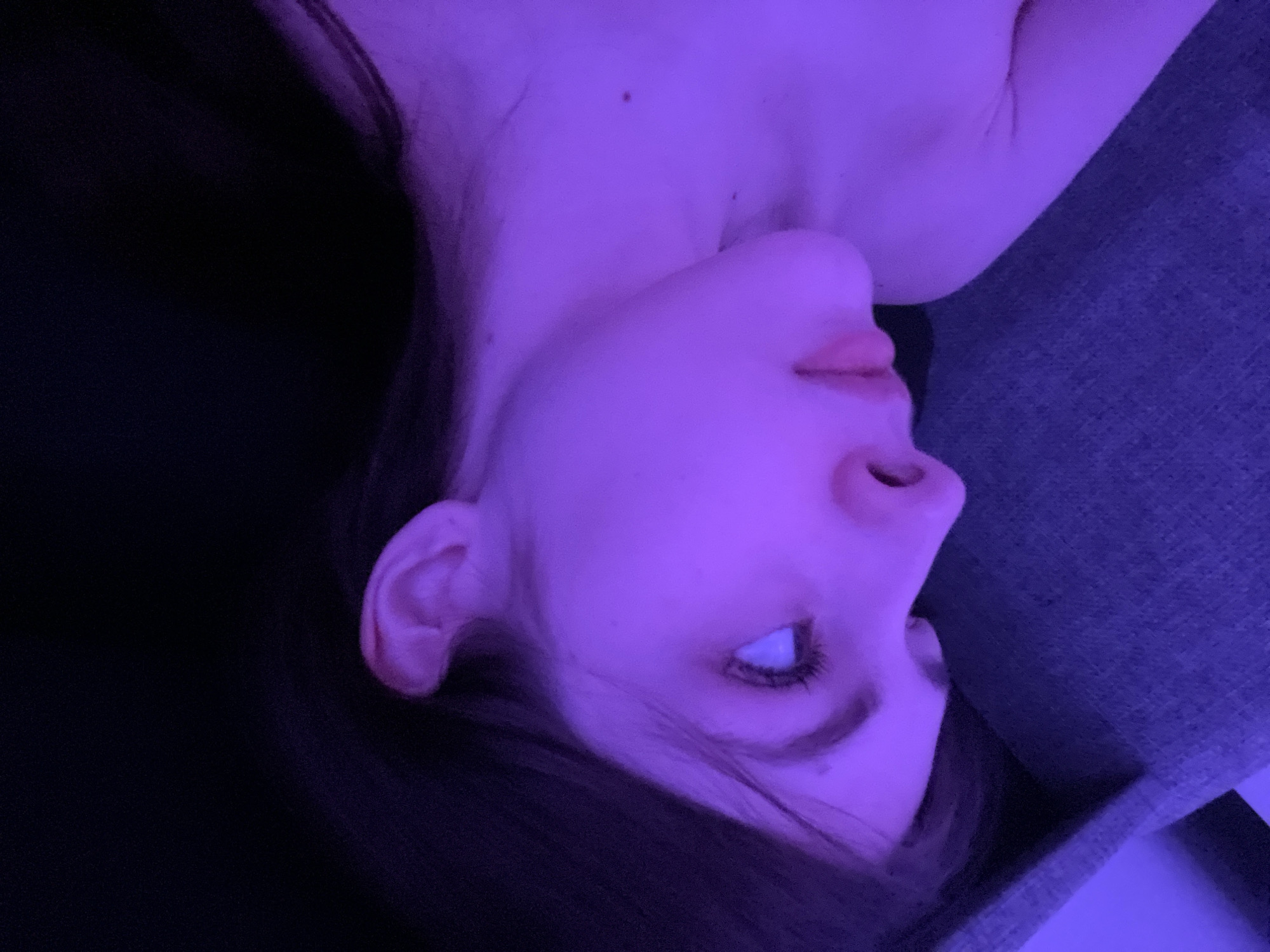 The image shows a close-up of a person's face and upper chest, illuminated by a purple light. The person has fair skin, dark hair, and one eye is visible. The background is dark and indistinct on the left, while on the right, there is a textured surface, possibly fabric. There is no indication of the location being Saint Petersburg, Russia.