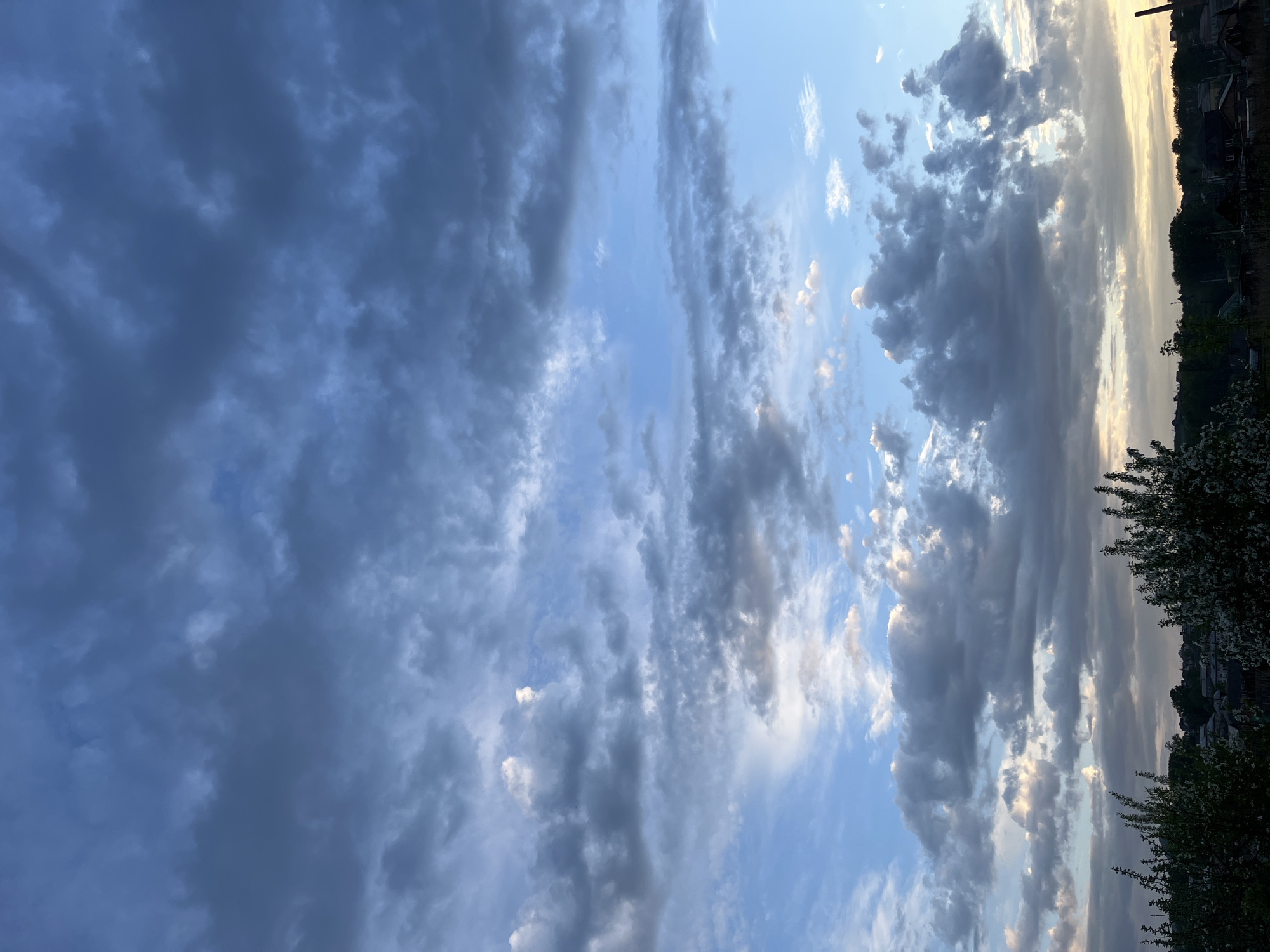 The image shows a sky filled with clouds, ranging from dark, ominous storm clouds on the left to lighter, more scattered clouds towards the right. The sky is predominantly blue, with patches of clear sky visible between the clouds. The photo seems to have been taken near Зыково, Russia, as evidenced by the presence of trees and rooftops along the top and right edges, typical of a populated area. The lighting suggests either early morning or late evening. There are no people or subjects immediately visible within the frame.